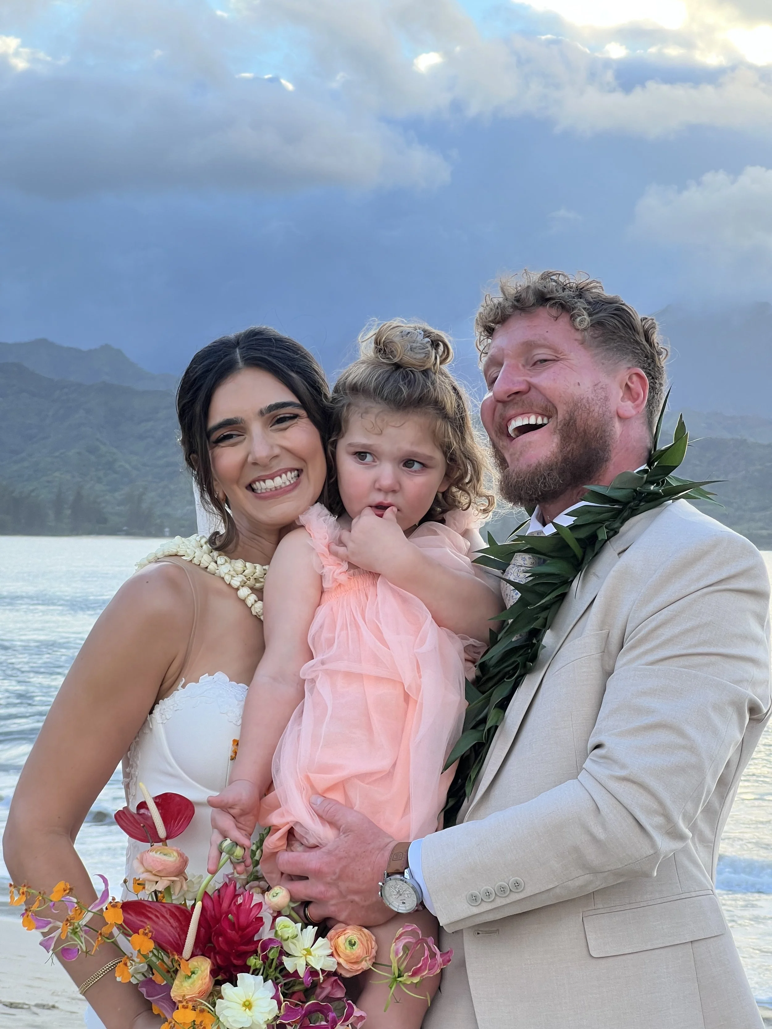 A couple dressed in wedding attire holding a young girl with curly hair and a pink dress, standing outdoors near water with mountains and cloudy sky in the background, surrounded by flowers.