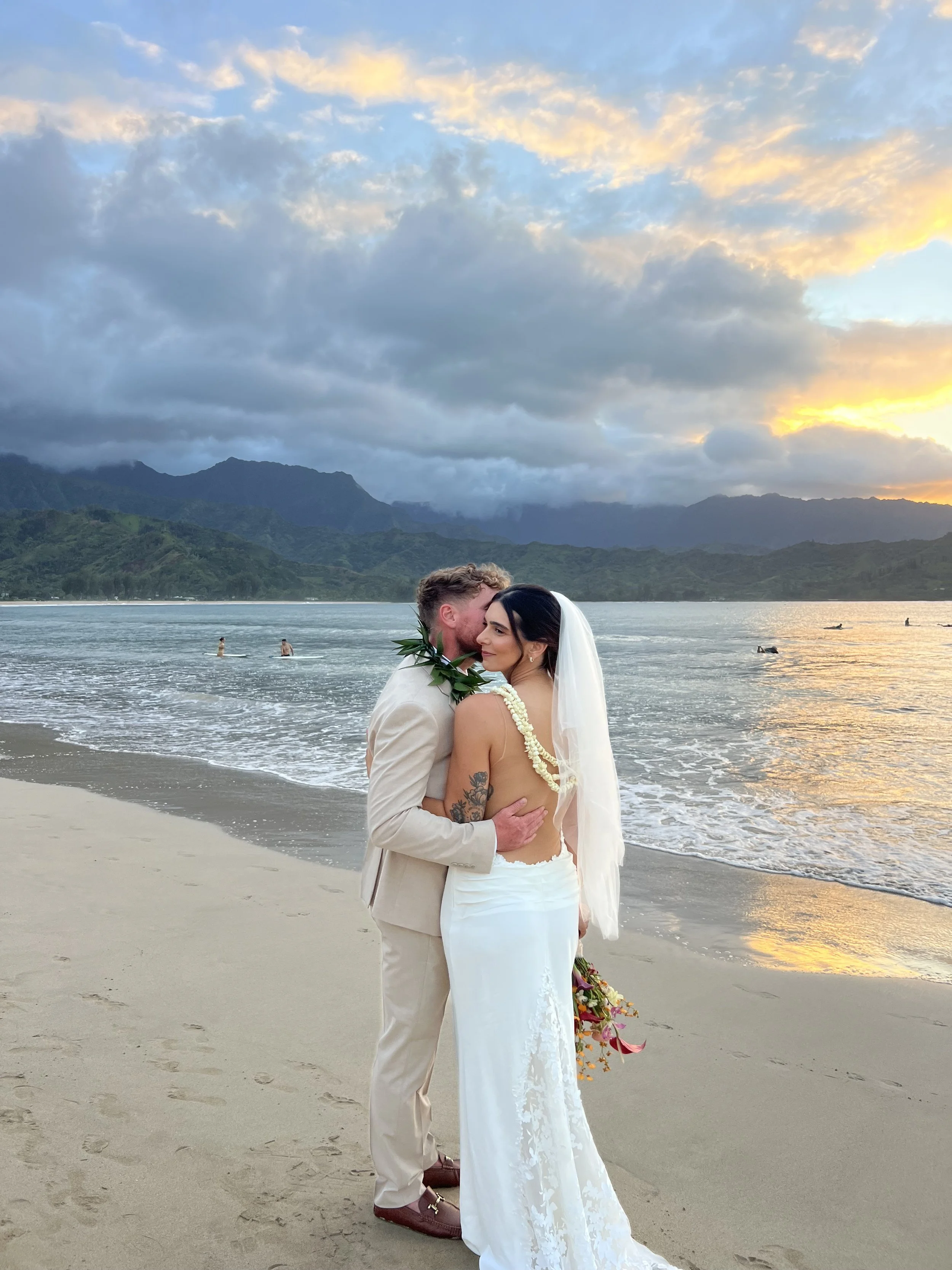 A couple dressed in wedding attire embracing on a beach at sunset, with mountains and clouds in the background.