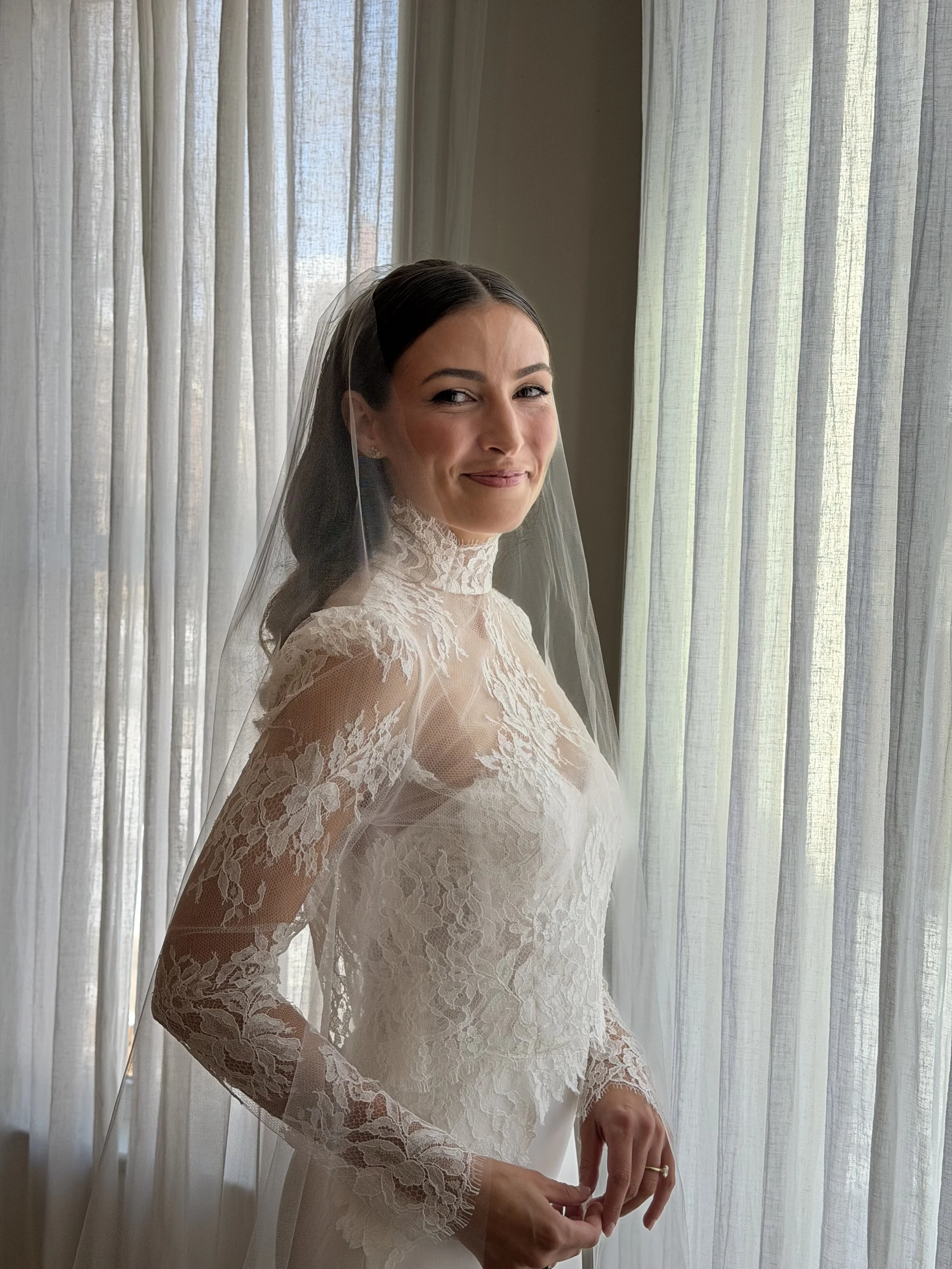 A woman dressed in a lace wedding gown with long sleeves, standing near sheer curtains with sunlight coming through, smiling softly, with dark hair styled back and wearing earrings and a wedding ring.