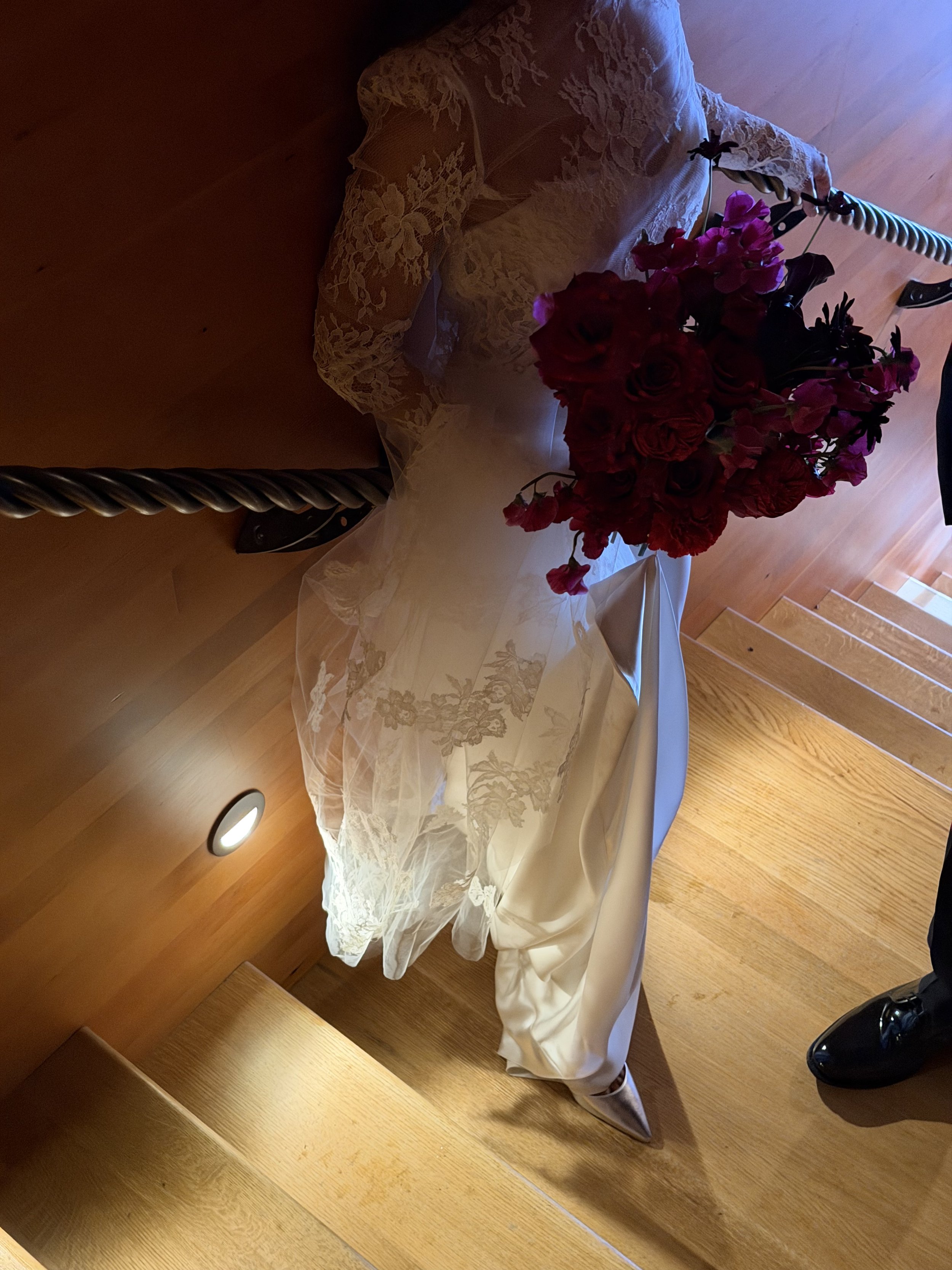 Close-up view of a small table with a lace tablecloth, decorated with a bouquet of red and pink flowers, next to a tied-up curtain and a pair of black shoes.