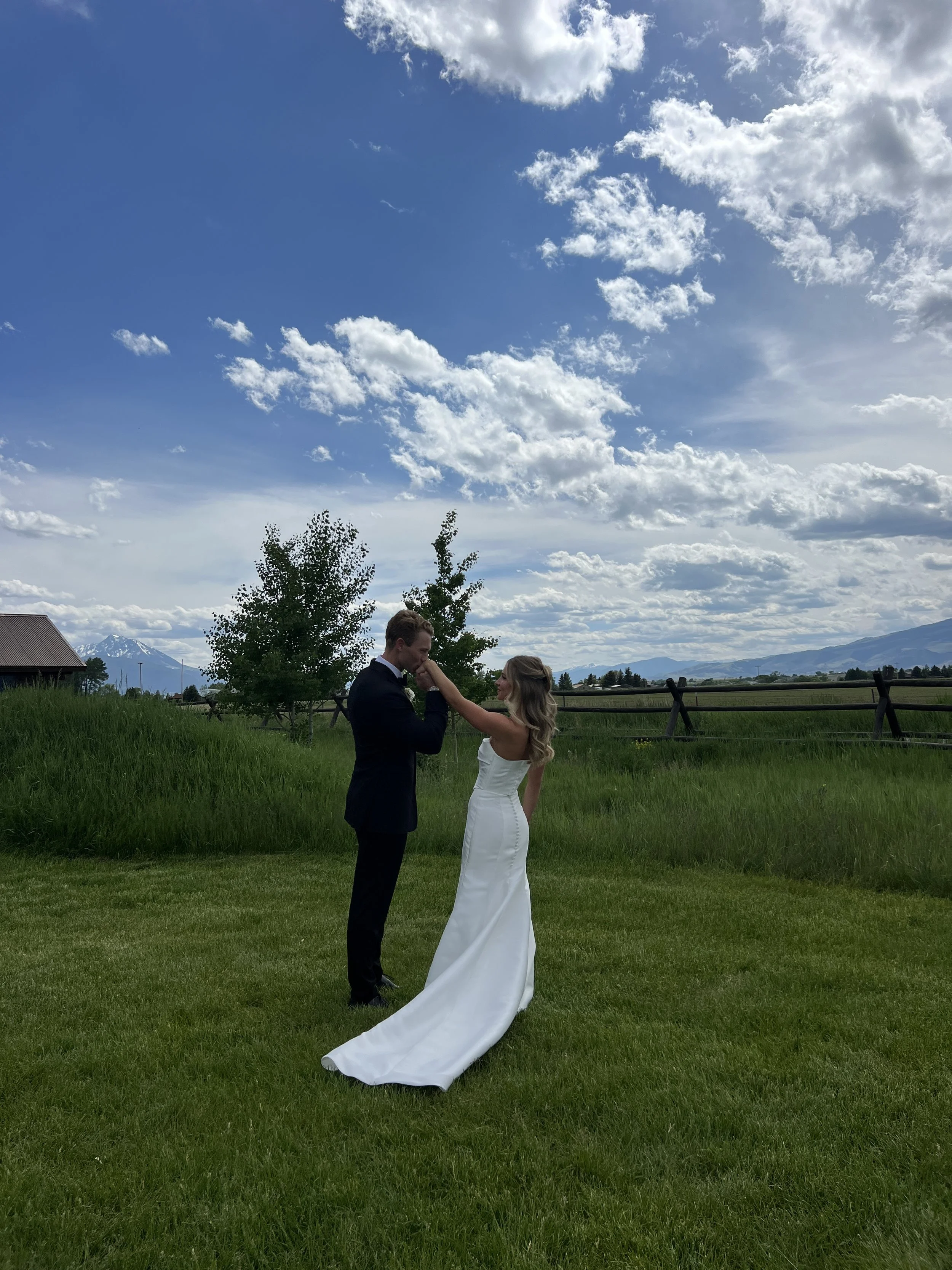 A newlywed couple holding hands and looking at each other outside on a grassy field with mountains in the background and a partly cloudy sky above.