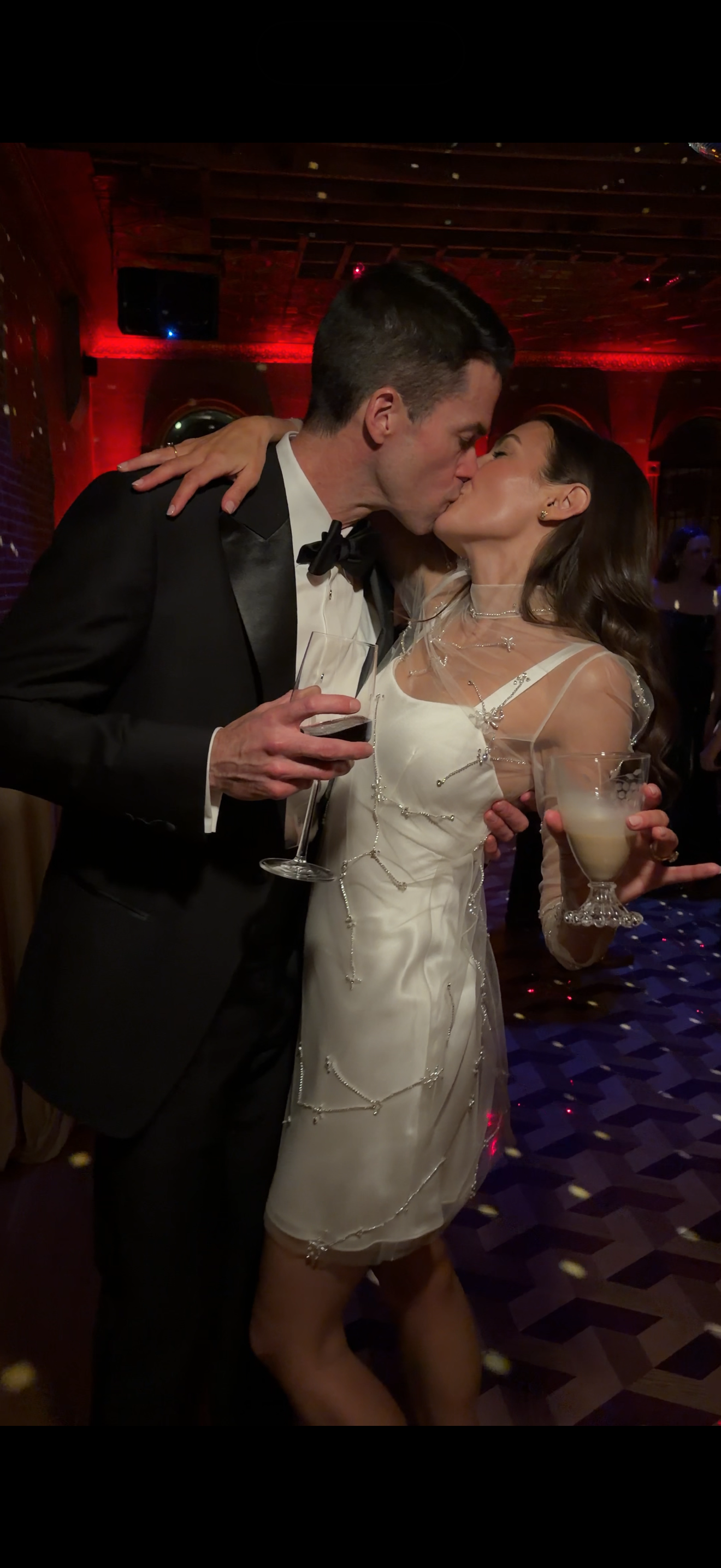 A man and woman kissing at a celebration, the woman is wearing a white dress with decorative beads, holding a drink, and the man is in a tuxedo holding a glass of red wine, in a dimly lit room with red lighting.