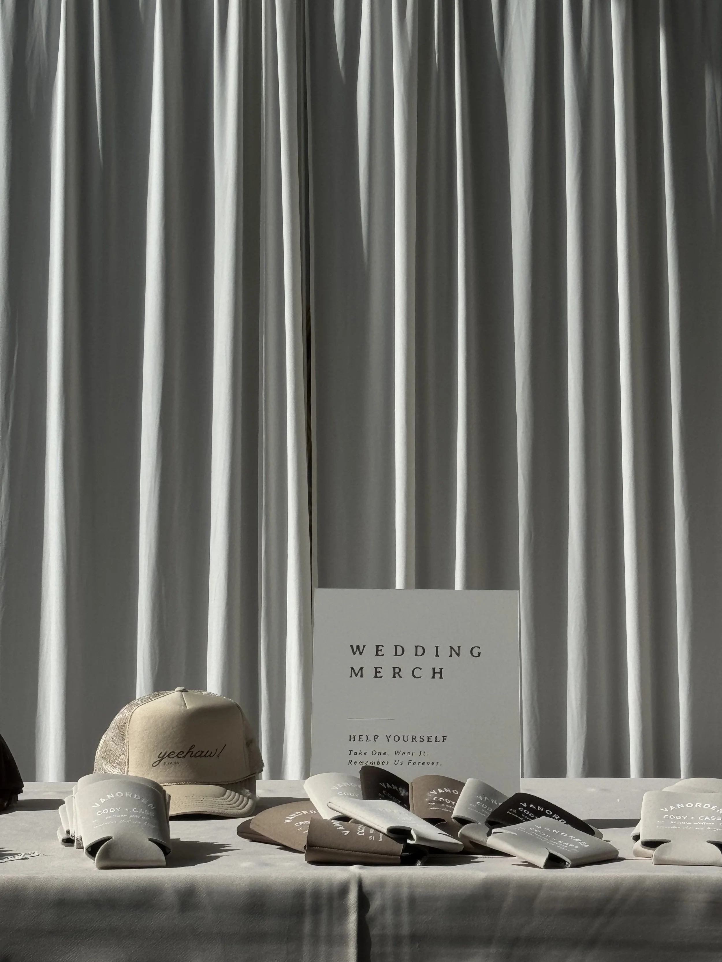 A display of wedding merchandise including beige and black caps, displayed on a table. Behind the table, there is a white board with black text reading 'Wedding Merch' and more text encouraging help, remembrance, and taking or wearing items.
