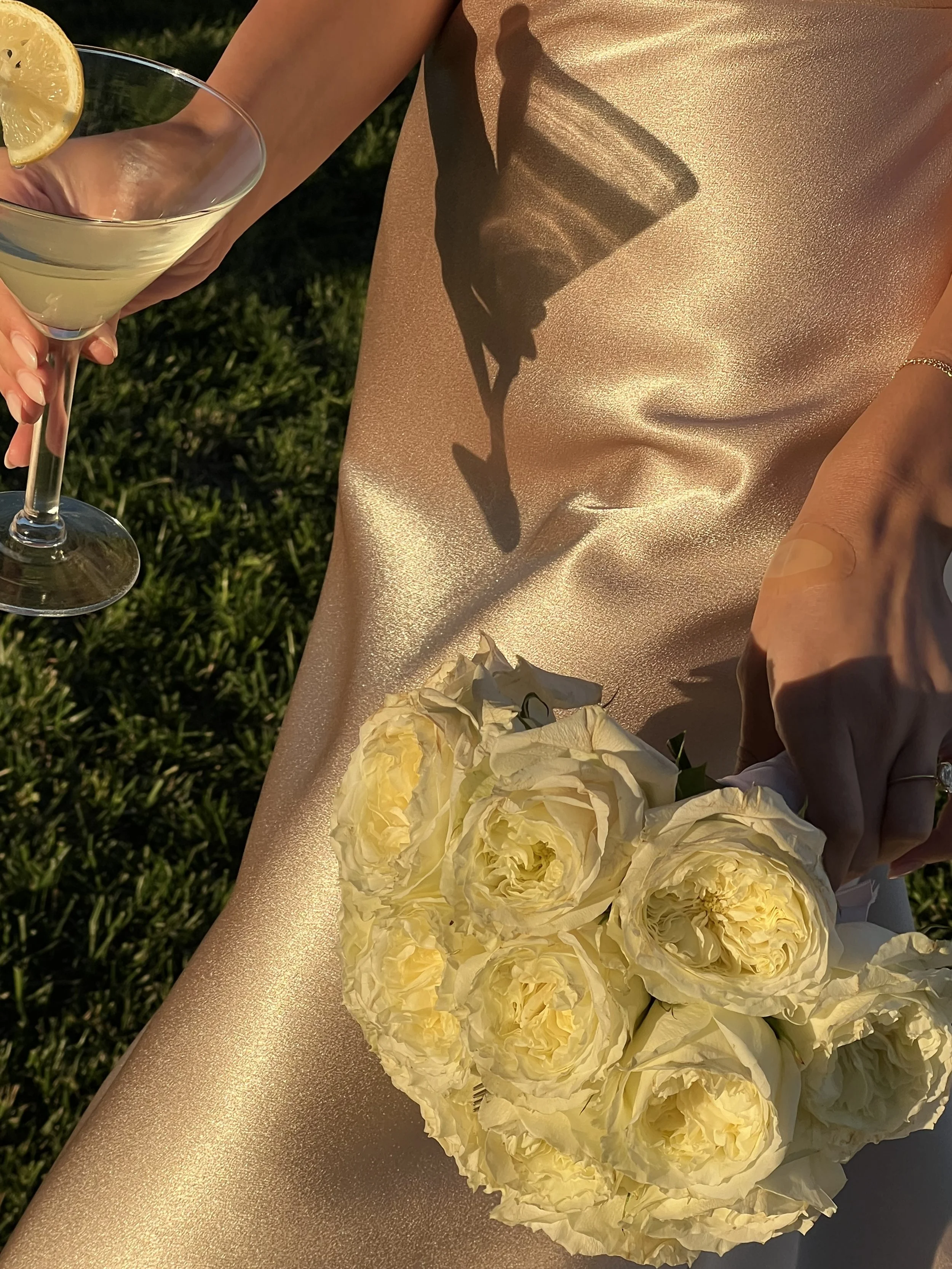 A woman's hand holding a bouquet of cream-colored peonies, with a gold-colored fabric underneath, a glass of lemonade with a lemon wedge, and sunlight casting shadows, including the shadow of a cup.