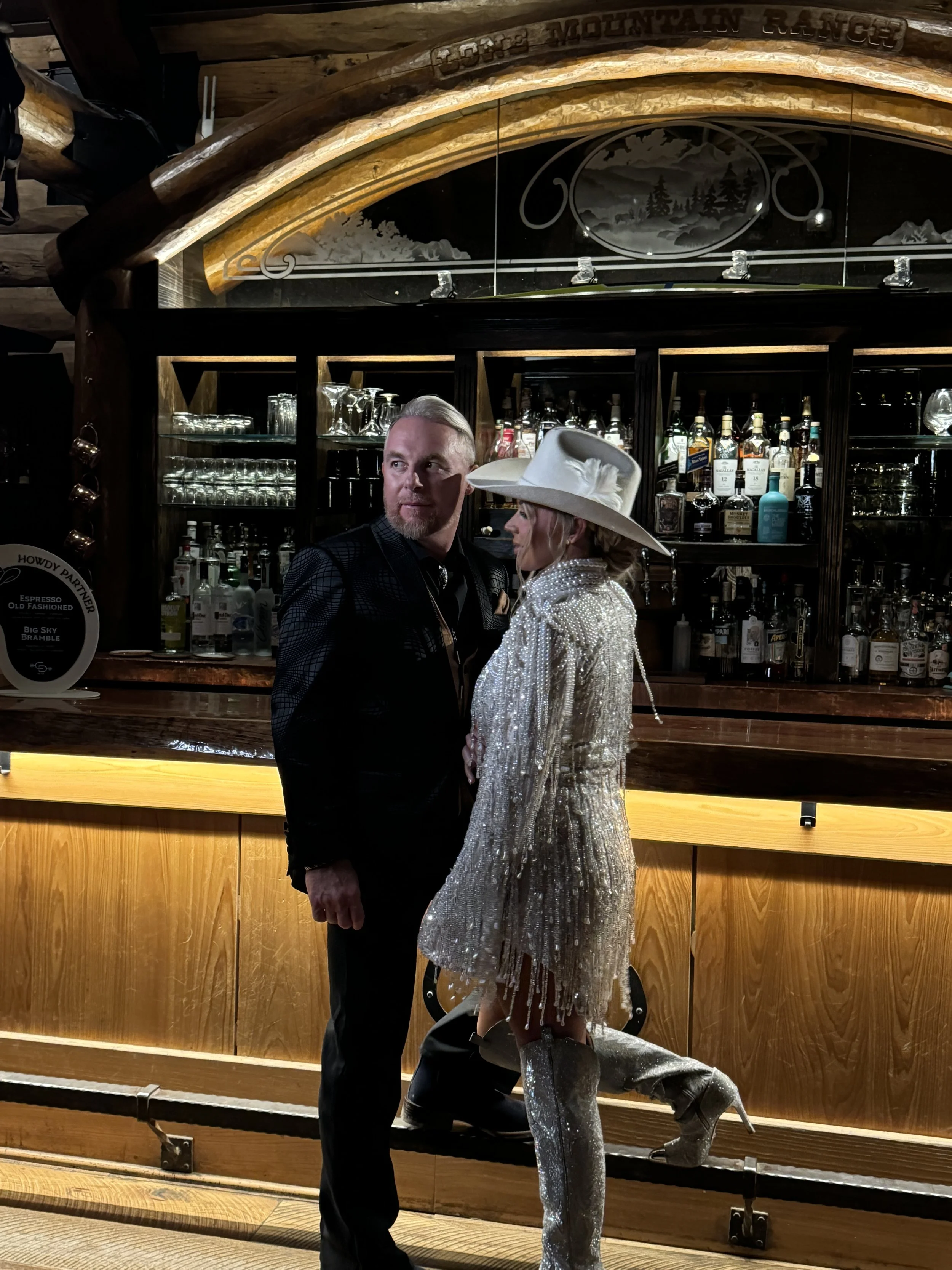 A man and woman standing indoors in front of a bar, with the woman wearing a large white hat with a feather and a shiny, silver, textured dress, and the man dressed in dark clothing. Behind them is a wooden bar with bottles of alcohol and glassware o