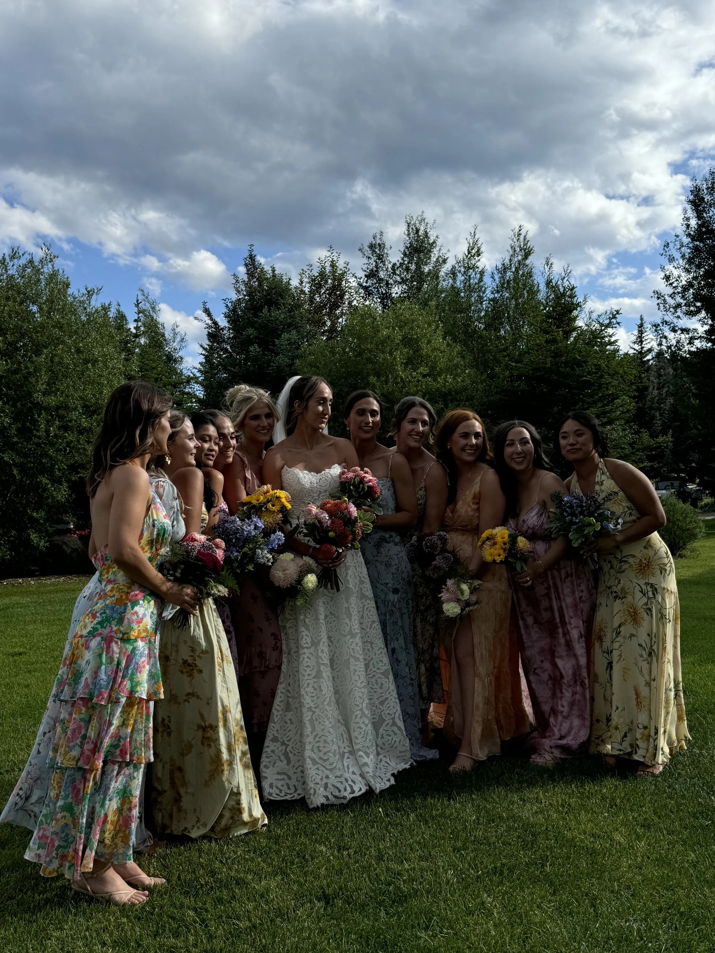 A group of women wearing colorful dresses, holding bouquets of flowers, standing outdoors on grass with trees and a cloudy sky in the background, celebrating a wedding.