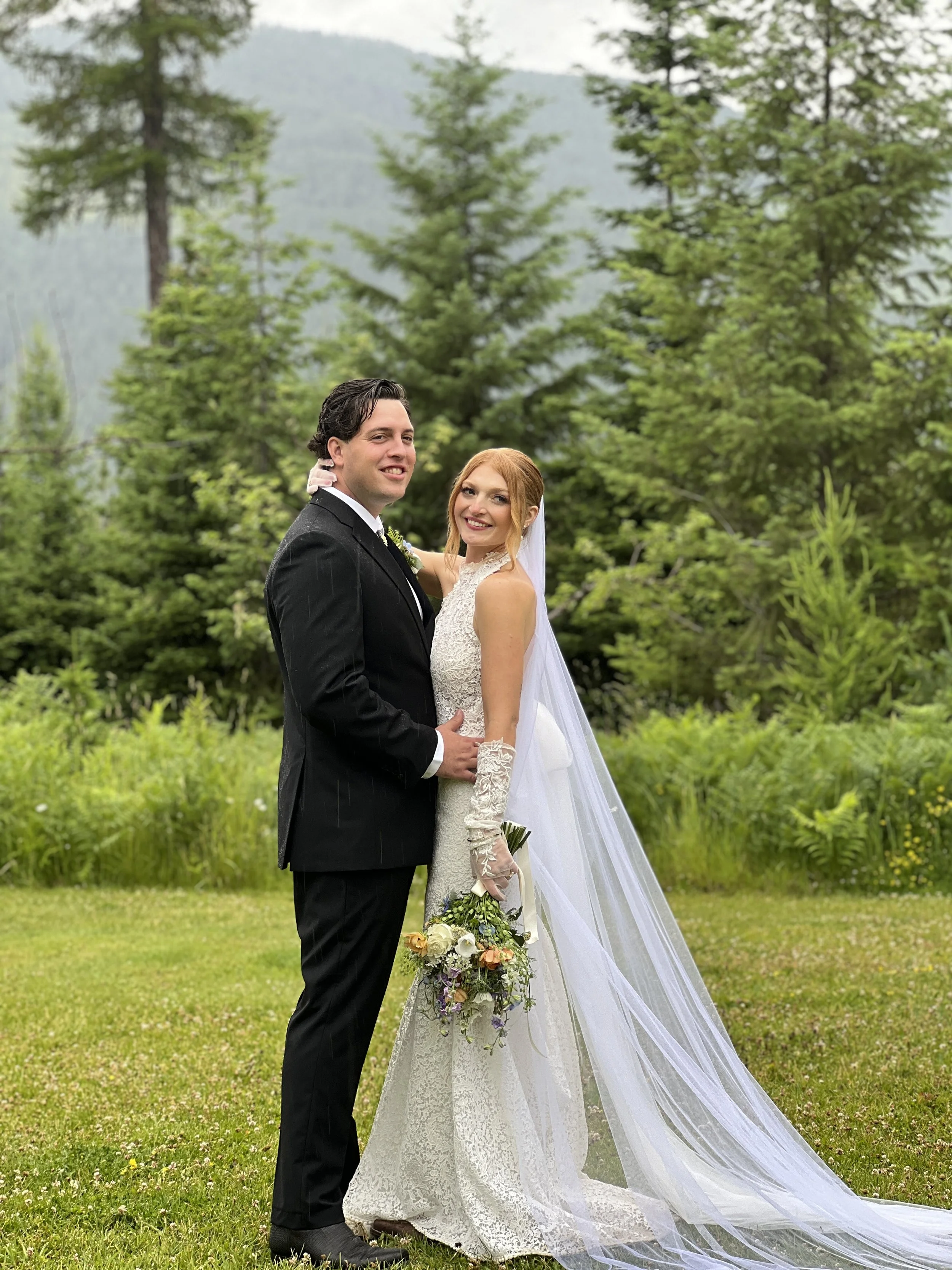A newlywed couple standing on a grassy field, surrounded by lush green trees, with mountains in the background. The groom is wearing a black suit and tie, and the bride is in a white lace wedding dress holding a floral bouquet, both smiling at the ca