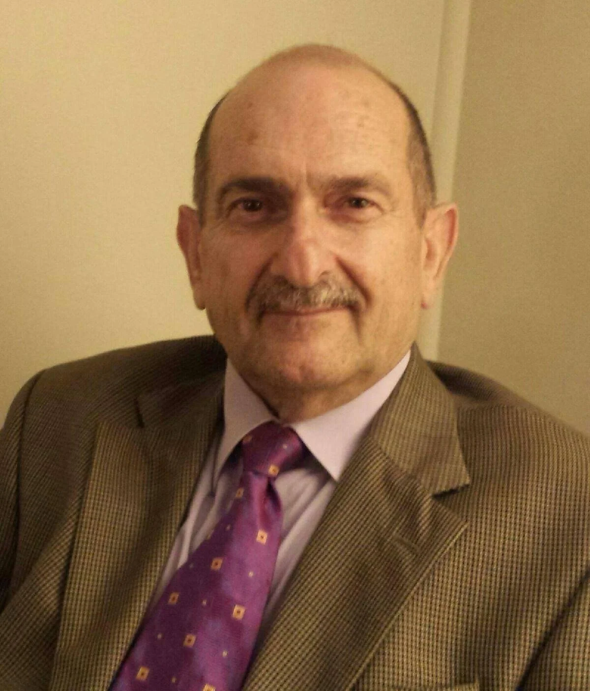 Portrait of an elderly man smiling, wearing a beige plaid blazer and a light blue collared shirt, with a lanyard around his neck.