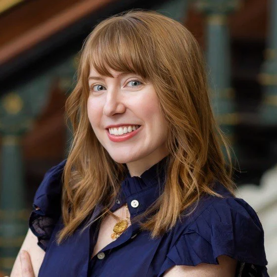 A woman in a dark navy -blue top with a light-colored striped tie, standing outdoors in front of modern apartment buildings, looking confidently at the camera.