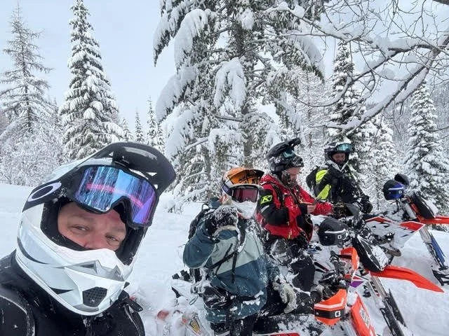 Four snowbike riders with their bikes in a snow-covered forest