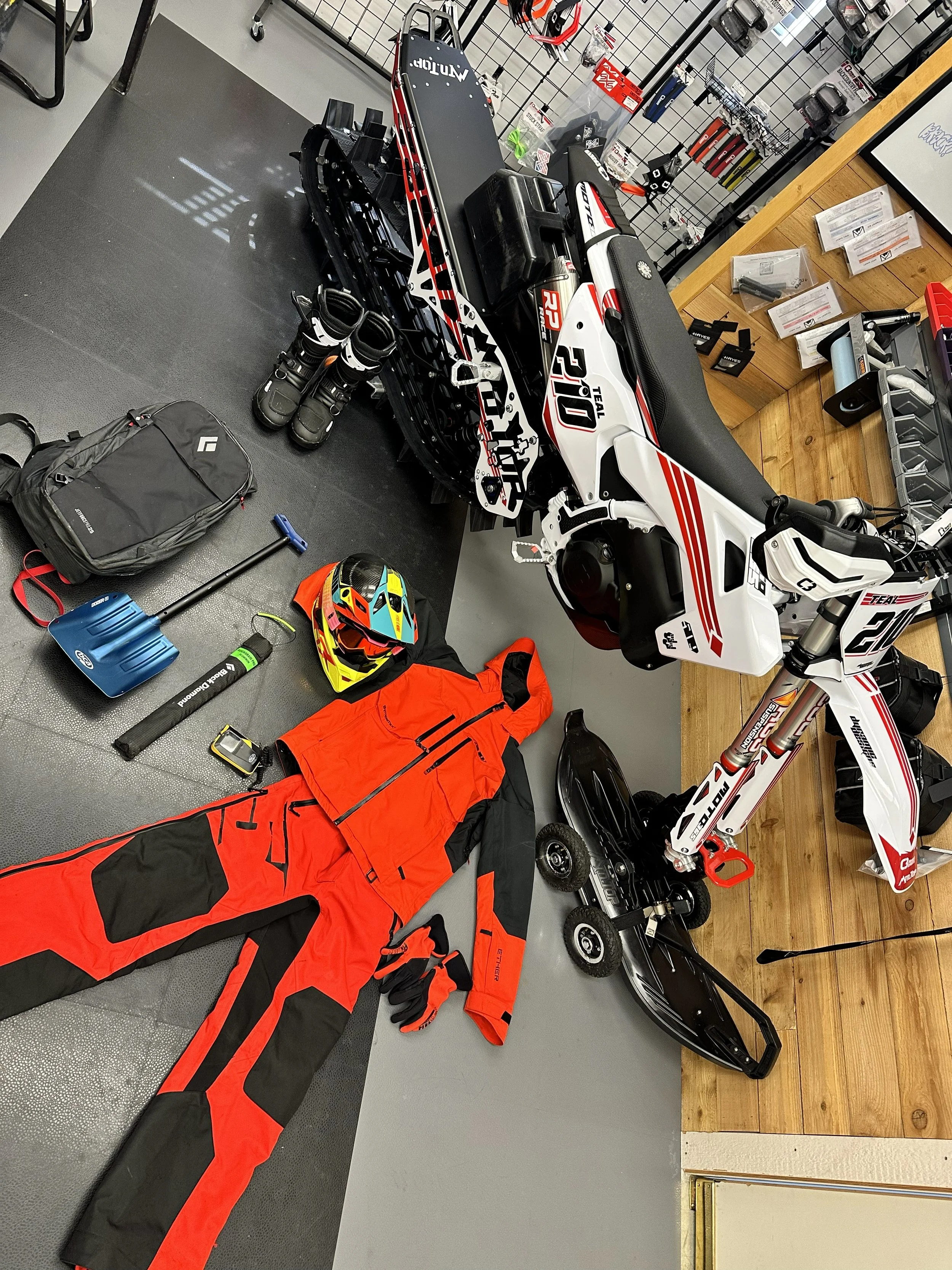 KTM dirt bike, snowbike kit, riding gear, and avalanche safety equipment laid out in the Moto365 shop.
