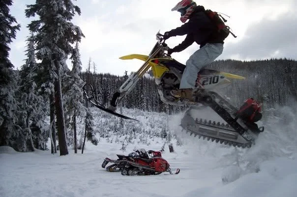 Early snowbike jumping beside parked snowmobiles in the backcountry