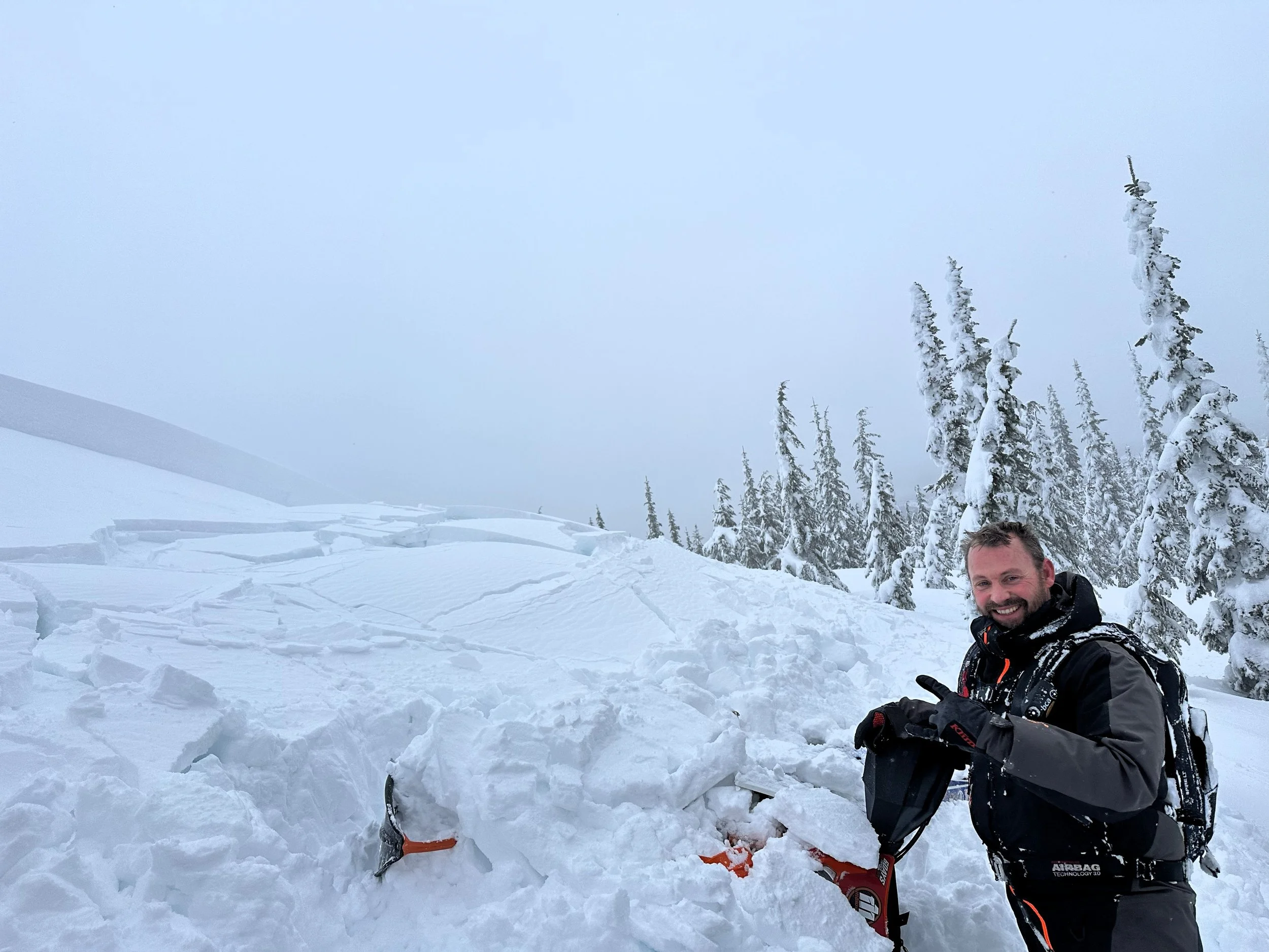 Rider holding a shovel beside a snowbike partly buried in deep snow, with snow-covered trees behind.