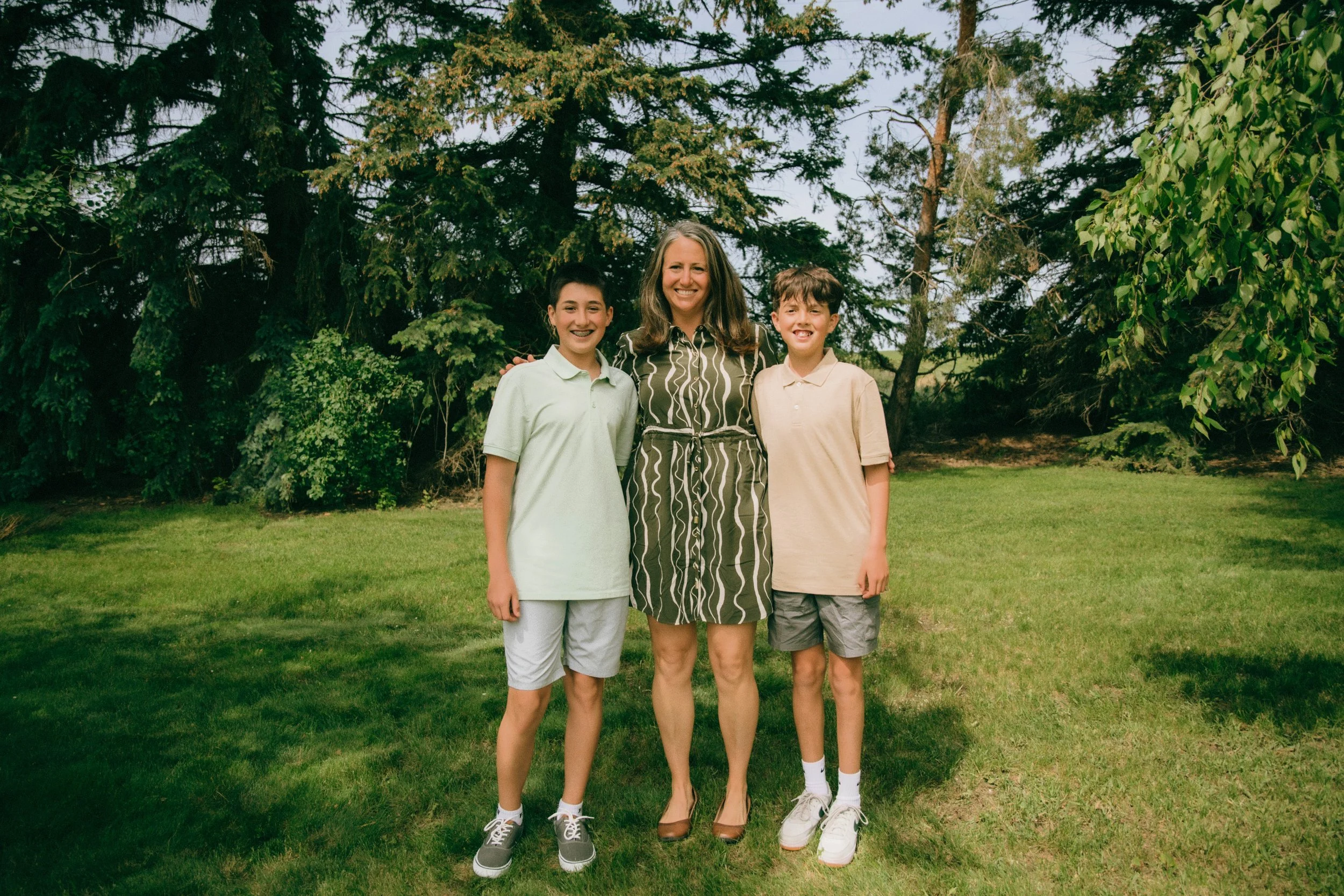 A woman and two boys smiling outdoors in a grassy area with trees in the background.