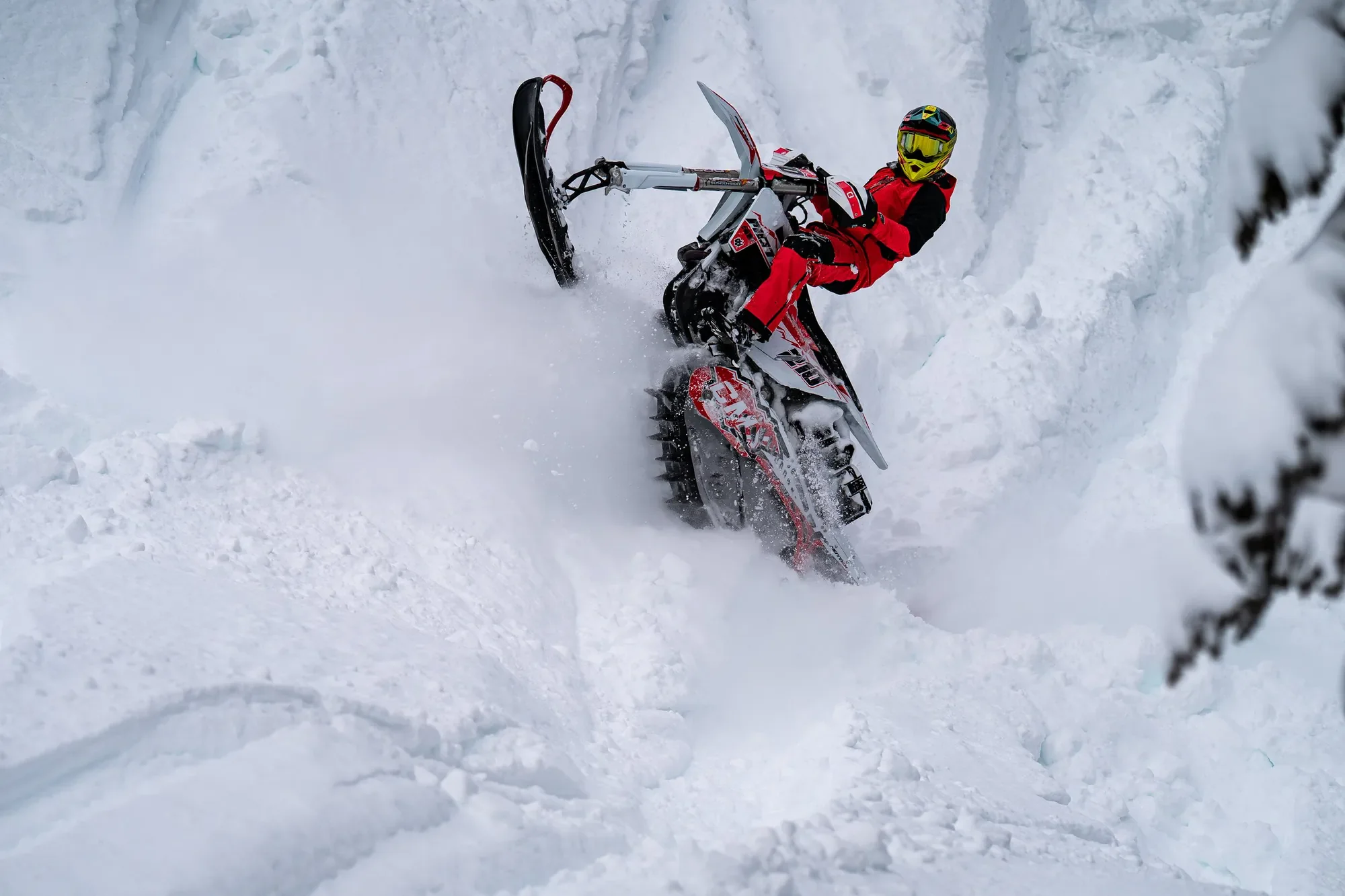 A person wearing a yellow helmet and red winter gear riding an Alpine snowbike on snow, leaning to the side while navigating through snow.