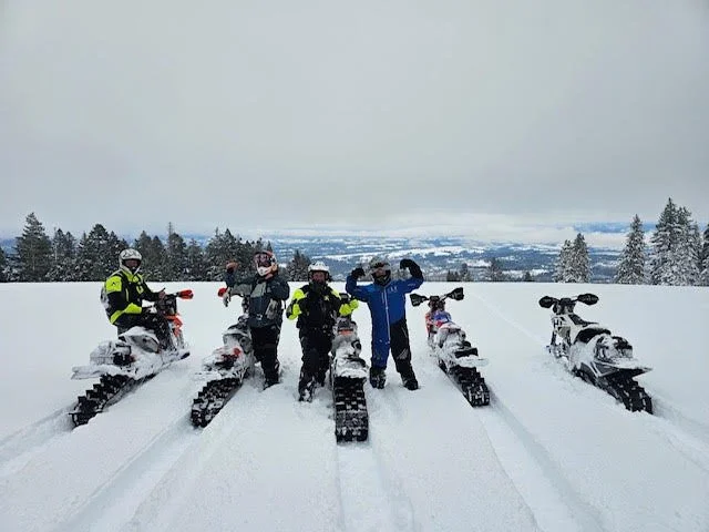 Five snowbike riders gathered with their bikes in a snowy field