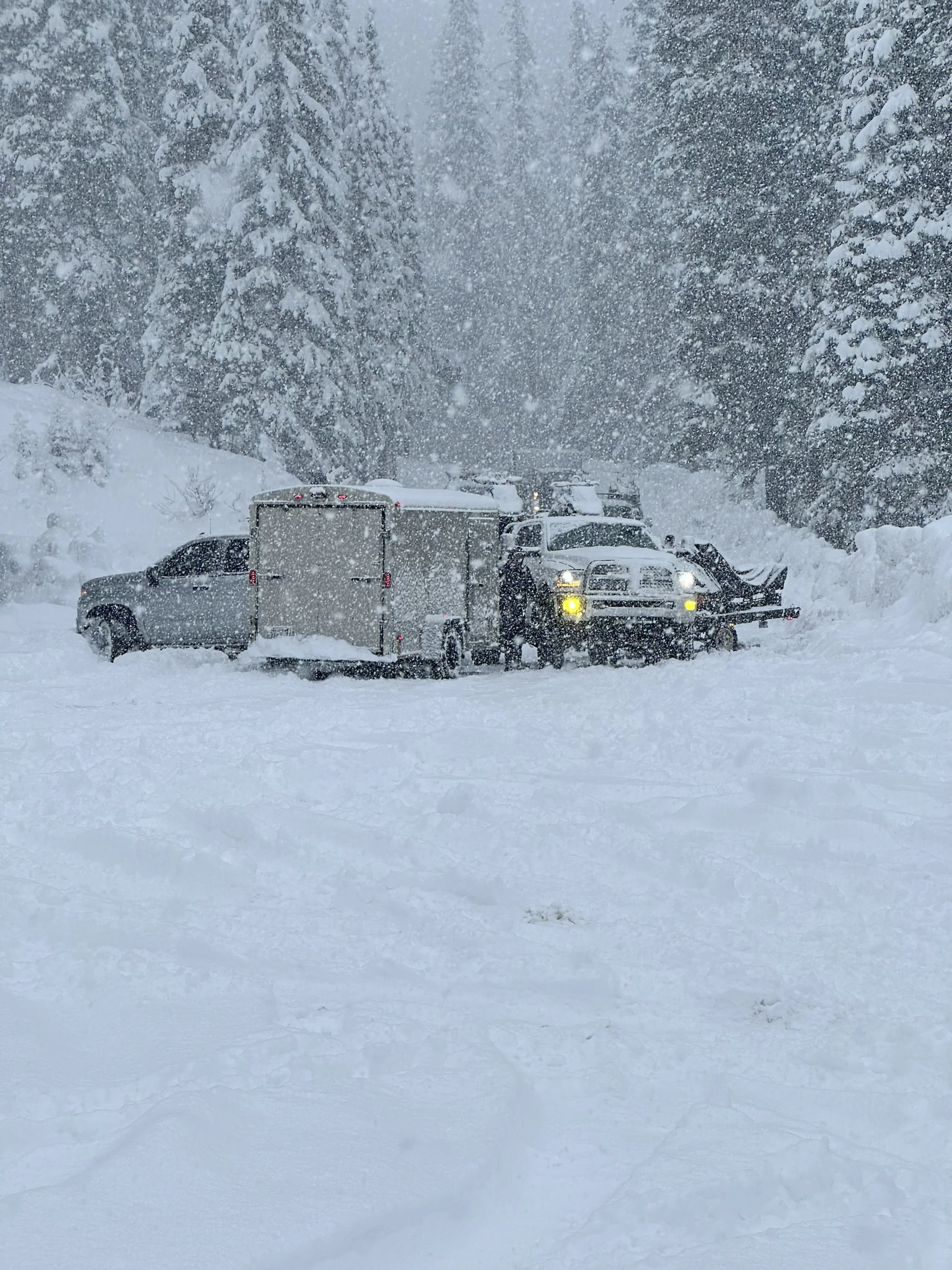 Pickup trucks and an enclosed trailer in a mountain parking area during heavy snowfall.