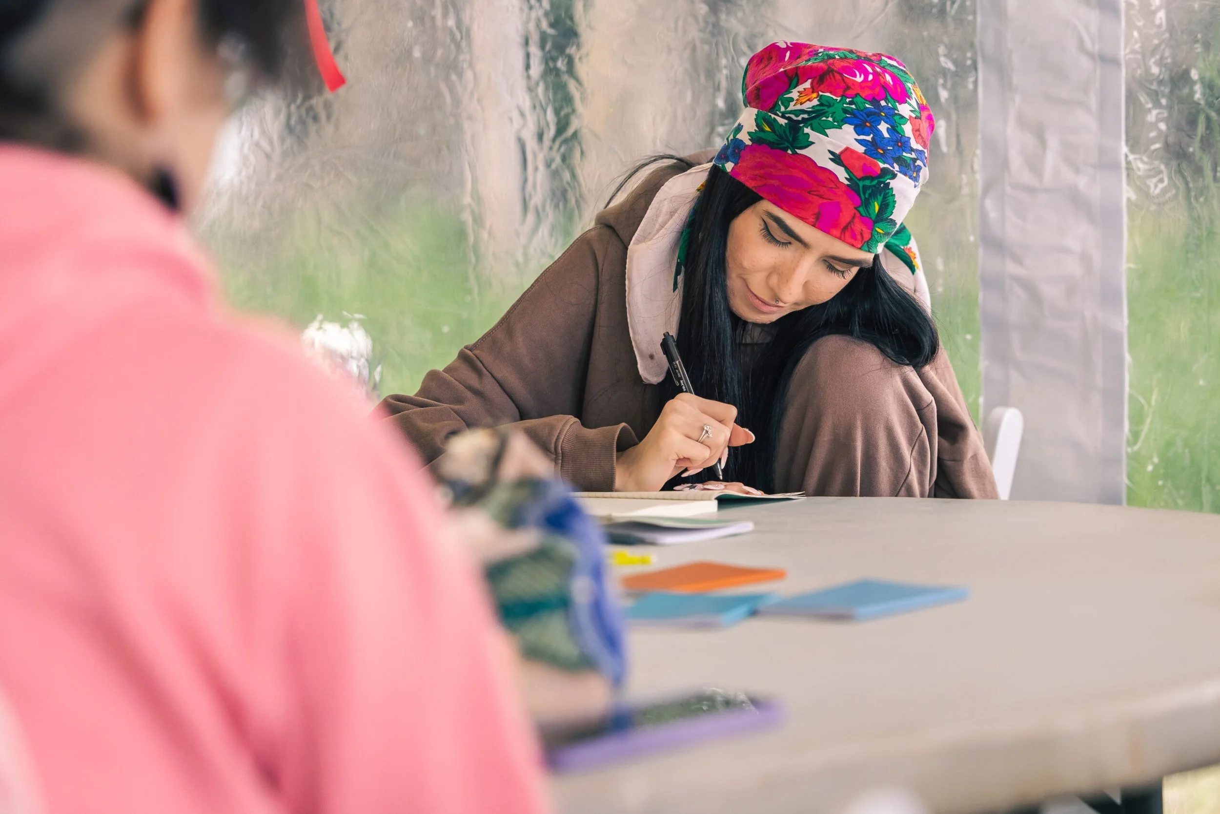 A woman wearing a colorful floral headscarf writing in a notebook at a table outdoors, with another person blurred in the foreground.