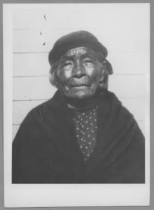 Black and white photograph of an elderly woman wearing a headscarf and a polka dot blouse, standing in front of a wooden wall.