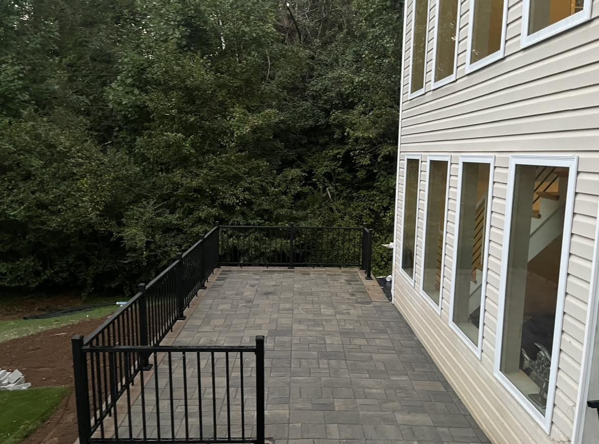 Outdoor patio with stone tiles and black railing adjacent to a beige-sided house with large windows, surrounded by dense trees.