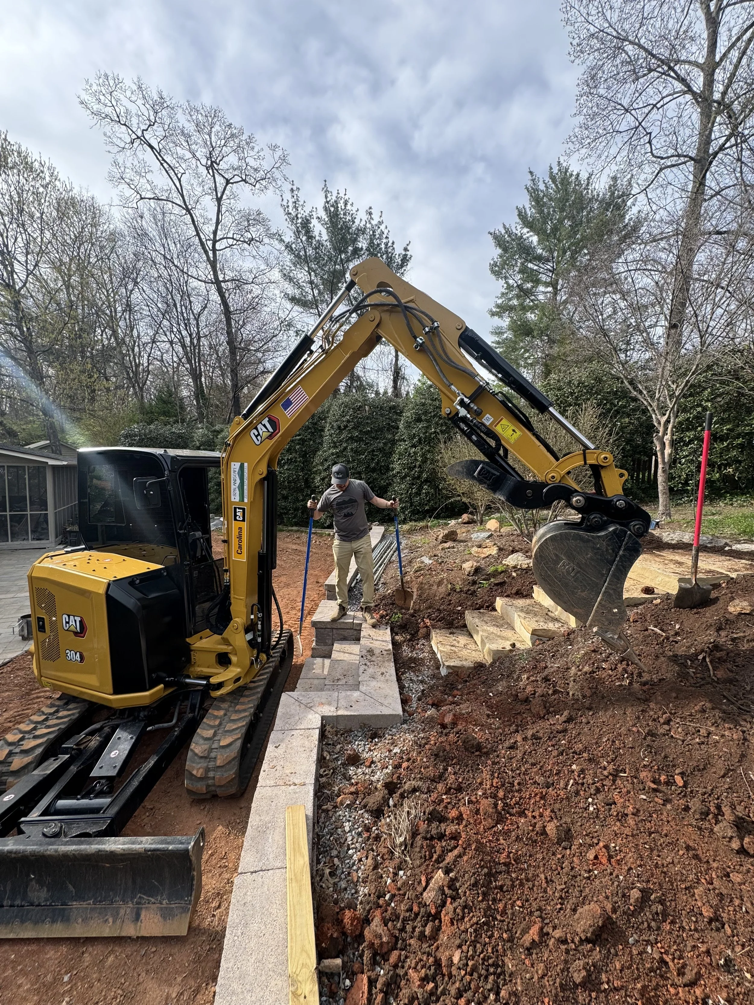 Yellow mini excavator digging in a garden area with a person nearby, surrounded by soil and trees.
