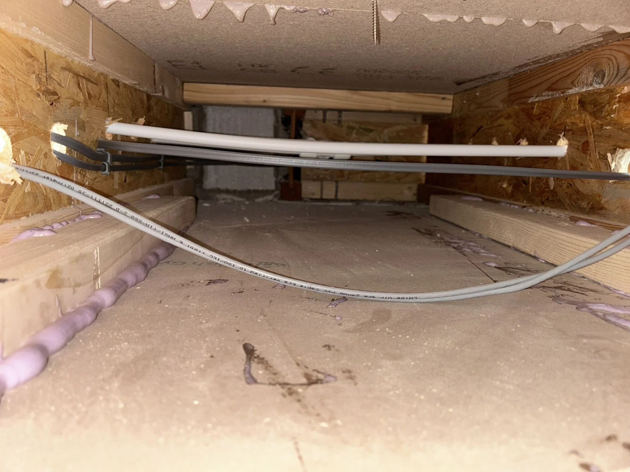 View of an unfinished basement or crawl space showing electrical wiring and plumbing pipes installed along the wooden framing and plywood floor.
