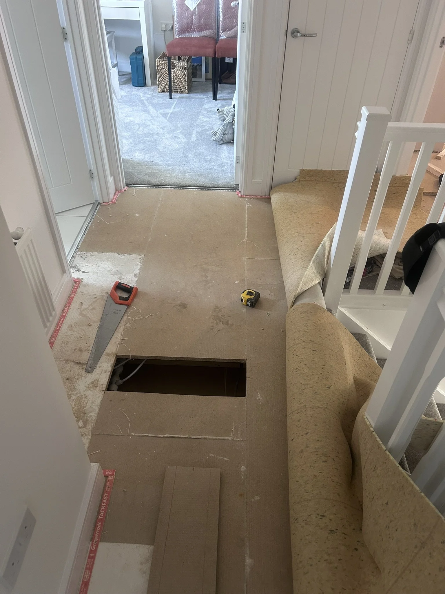 View of a room under renovation with the floor partially removed, exposing plumbing and a rectangular hole. Tools such as a saw, measuring tape, and a piece of cardboard are on the floor. There is a staircase with a rolled-up carpet and a retaining wall on the right side, leading to a lower level. In the background, a doorway reveals another room with chairs, a stuffed animal, and various household items.