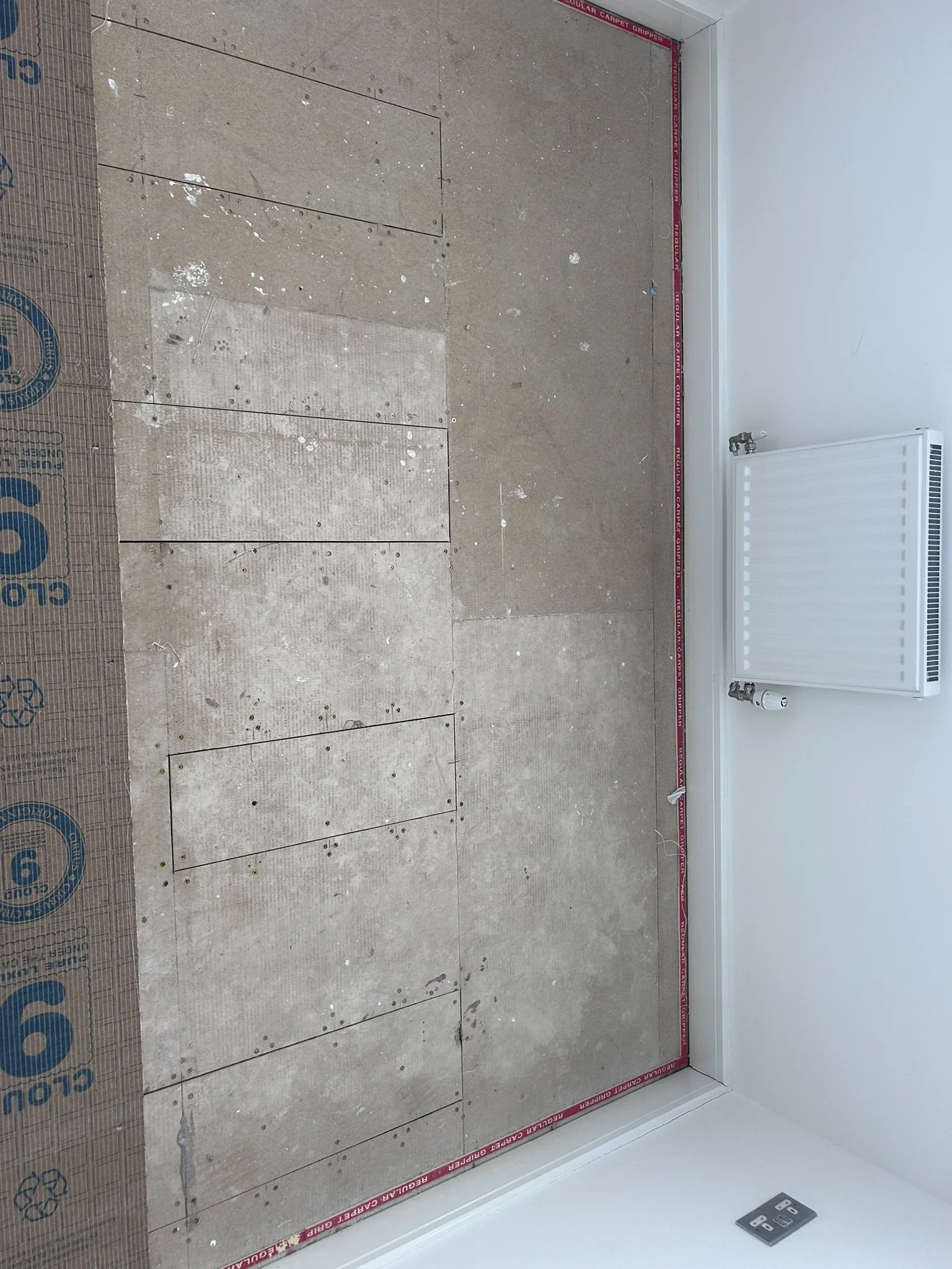 A room under renovation with a floor made of wooden planks, partially covered with construction material, and a white radiator mounted on the wall, with a power outlet nearby.