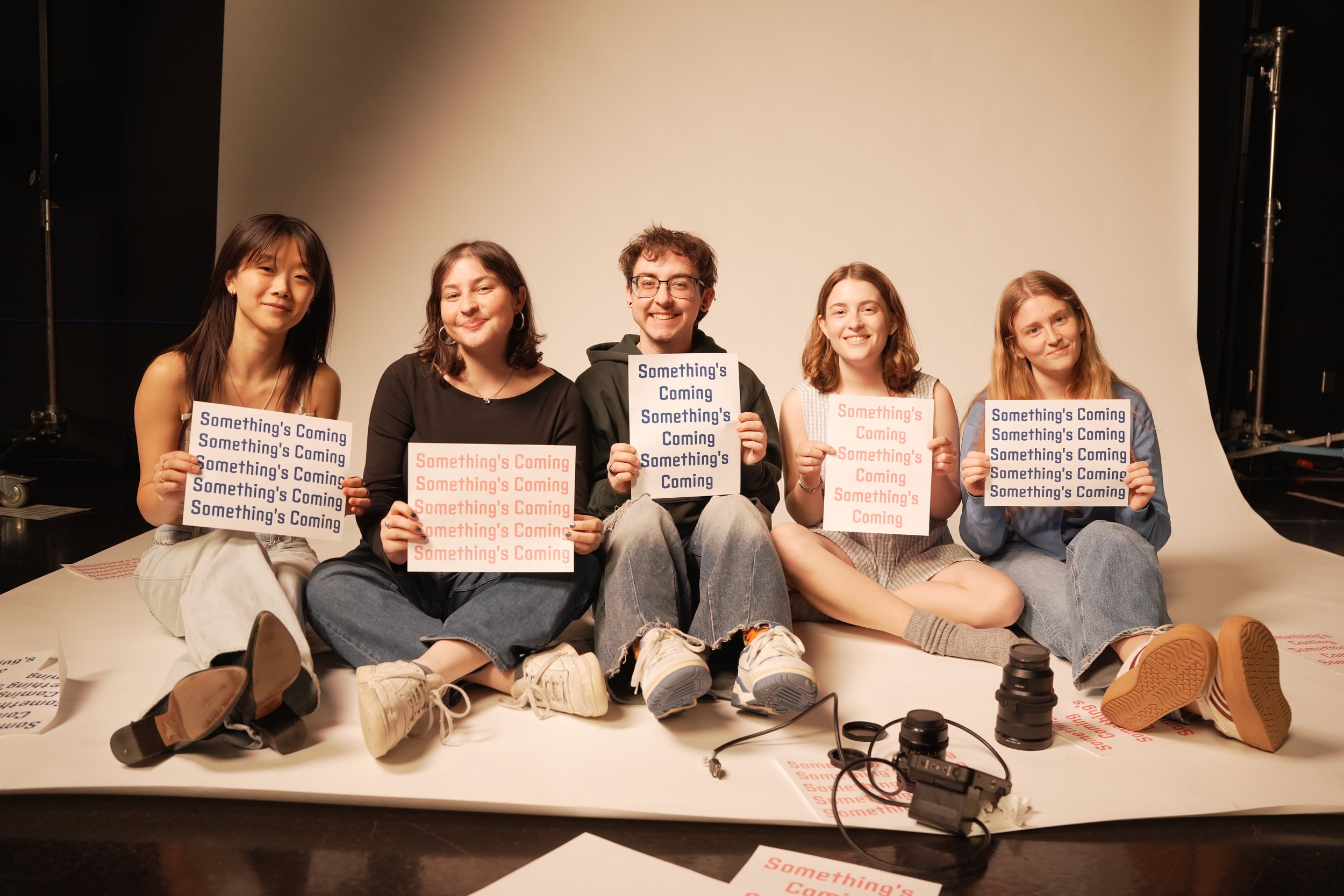 Five young people sitting on a white background with their legs stretched out, holding signs that read 'Something's Coming'. They are smiling and appear to be in a photo studio, with cameras and lighting equipment nearby.