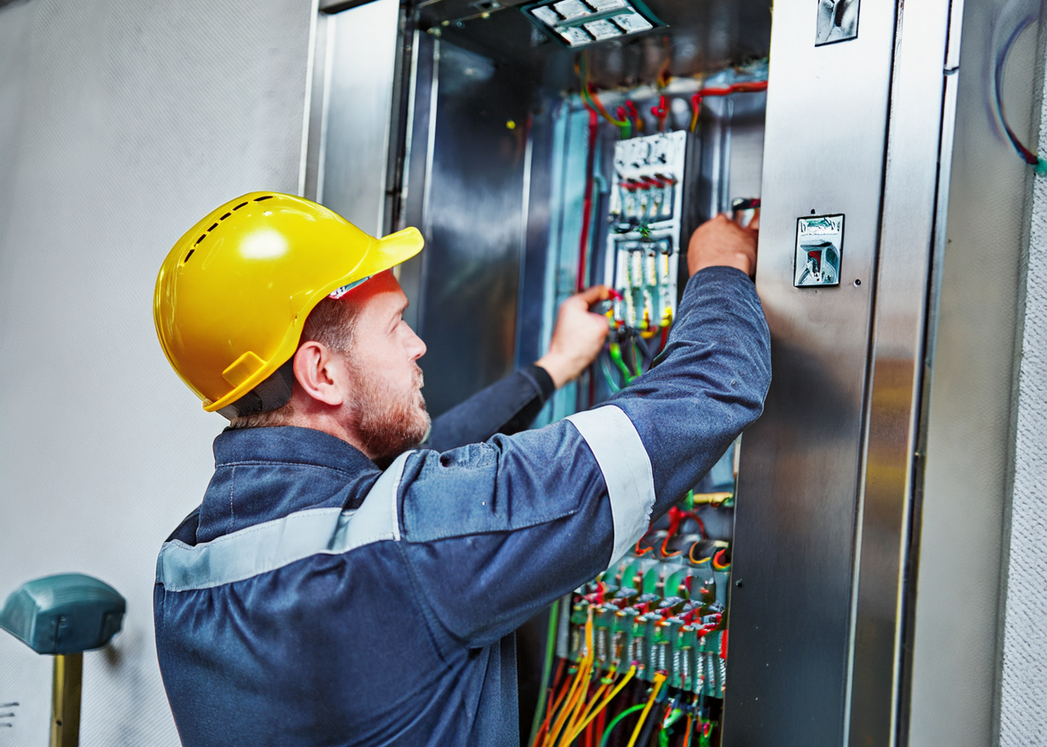A technician wearing a yellow safety helmet and a blue work uniform working on an electrical panel with colorful wires.