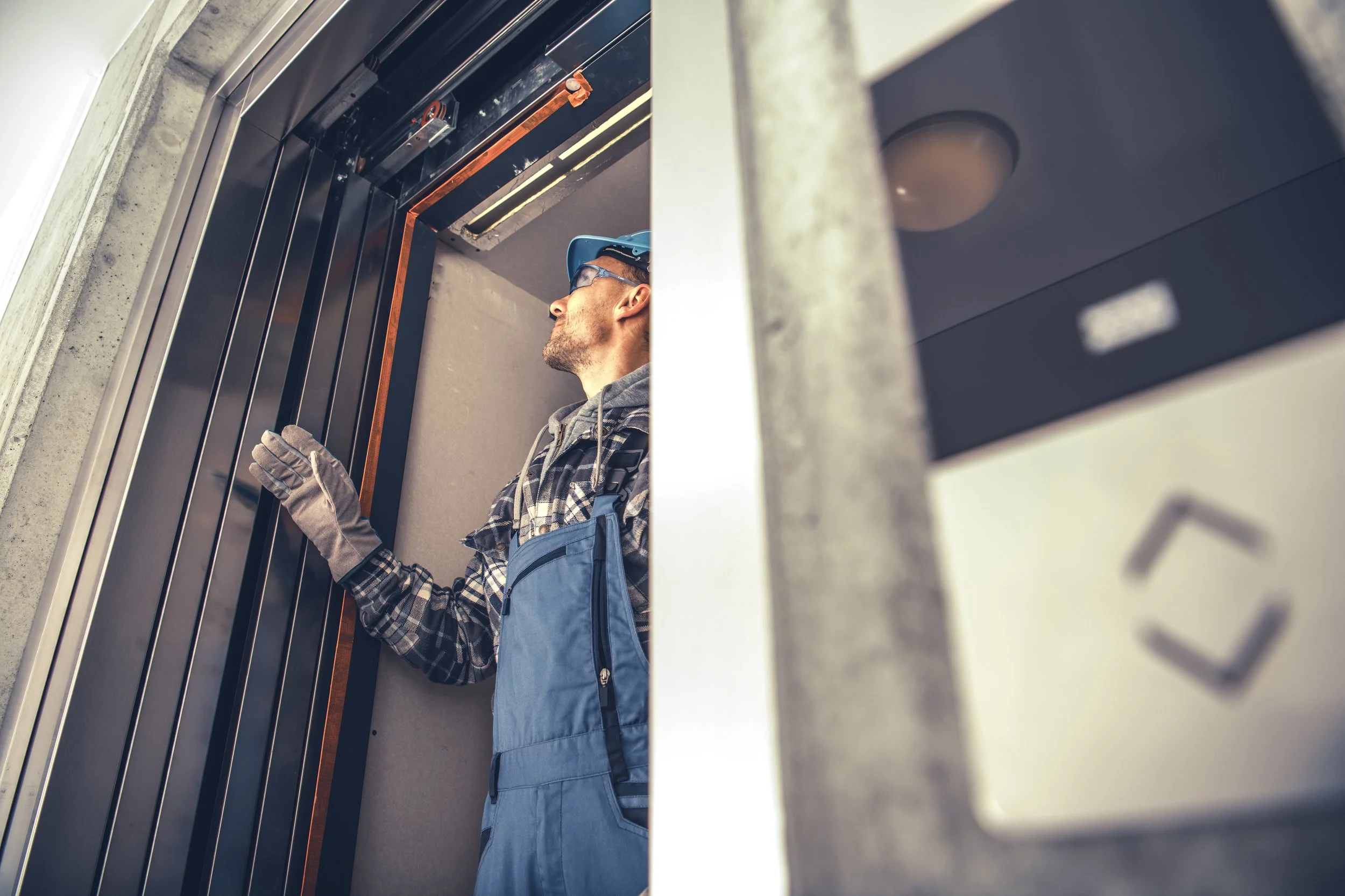 A man wearing safety glasses, gloves, and a plaid shirt is inspecting or working on an elevator shaft from inside an elevator car with the door open.