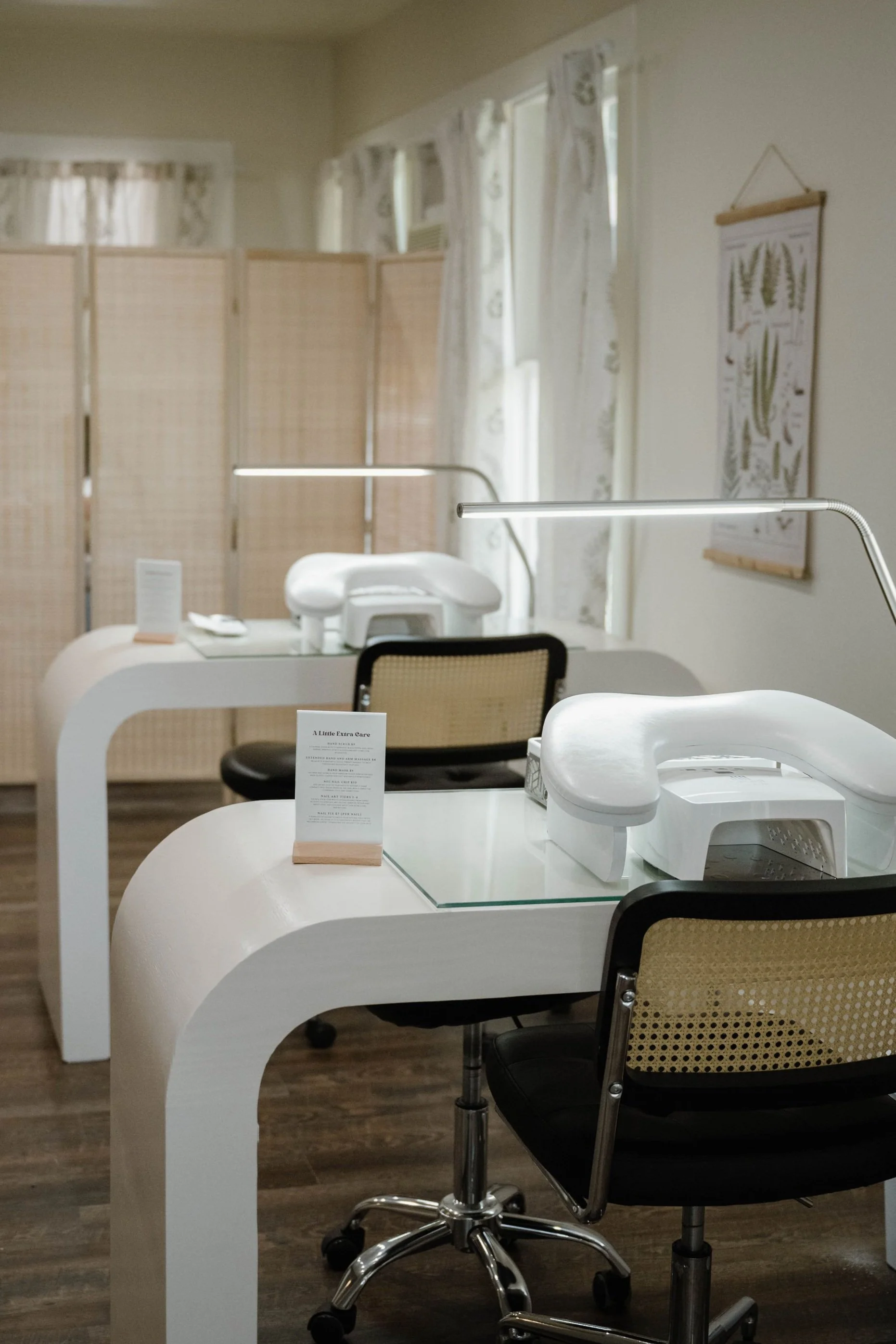 A room with two manicure or pedicure tables, each with a white armrest, a black and beige rolling chair, a small sign on each table, a glass top on one table, a printed poster of botanical illustrations on the wall, and a beige room divider in the background.