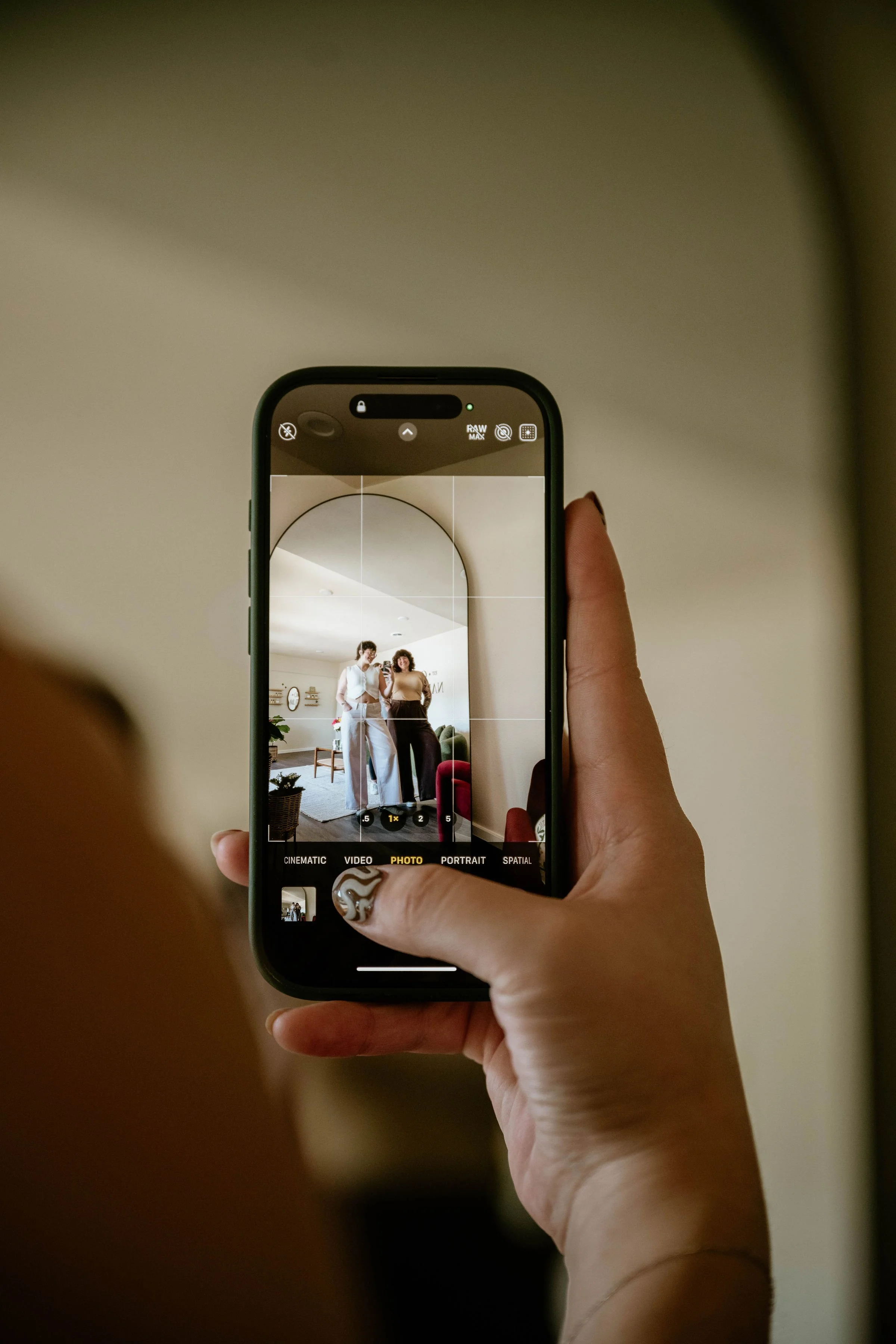 A person taking a photo of two women standing together inside a room, reflected in their smartphone camera.