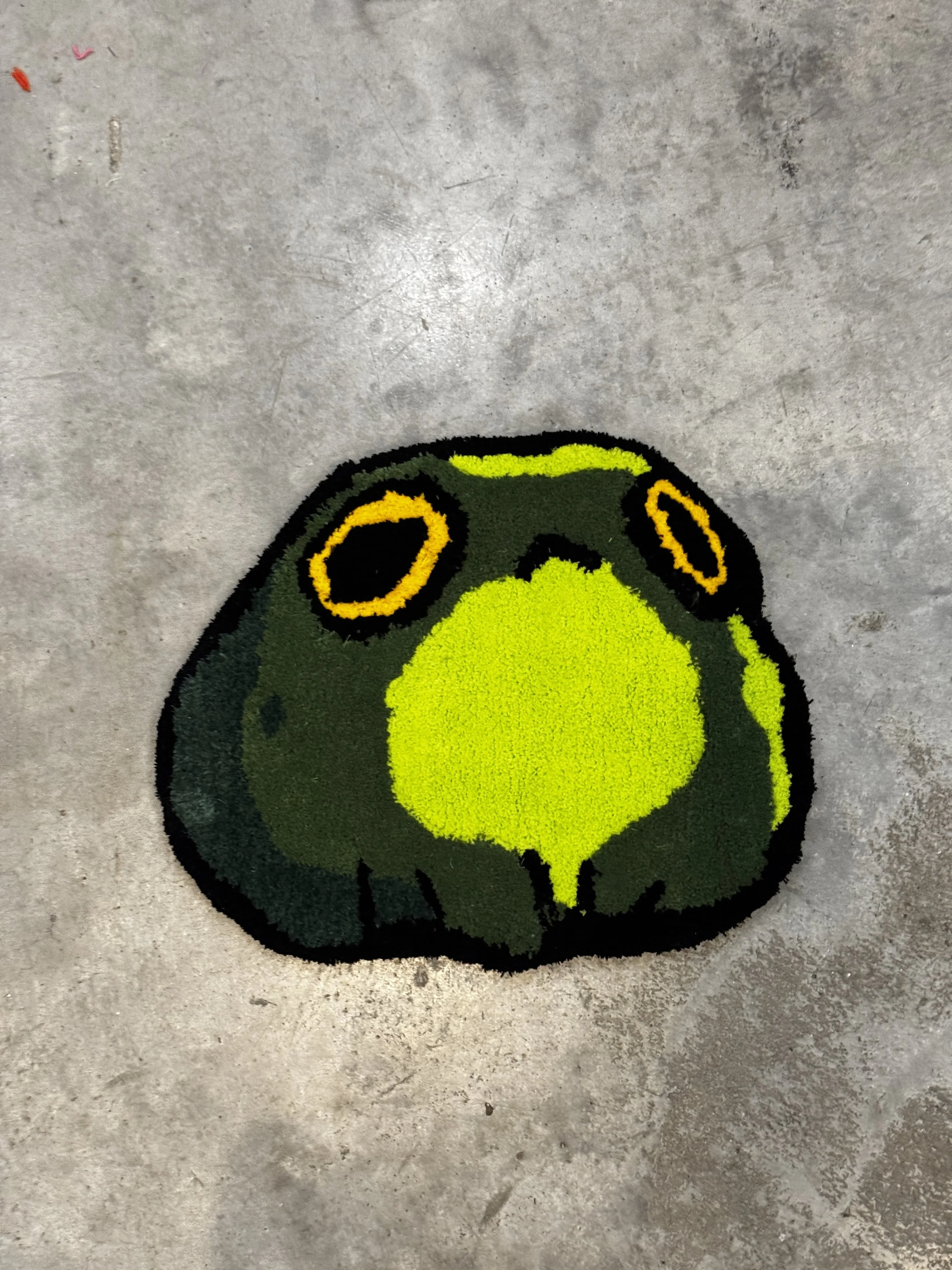 A green frog-shaped doormat with black and yellow accents on a gray concrete floor.