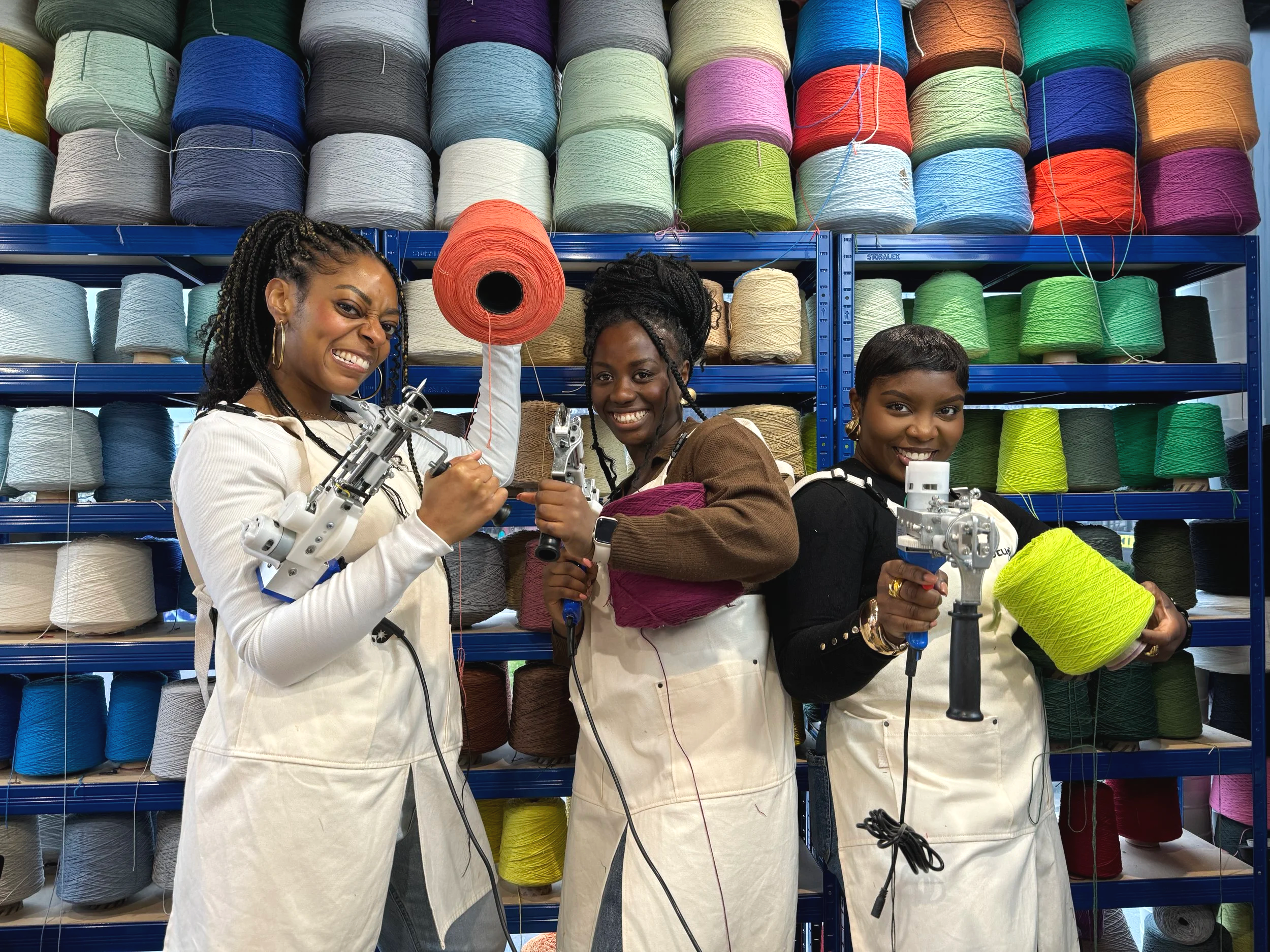 Three women in aprons working with a yarn winder in a yarn store filled with shelves of colorful yarn spools.