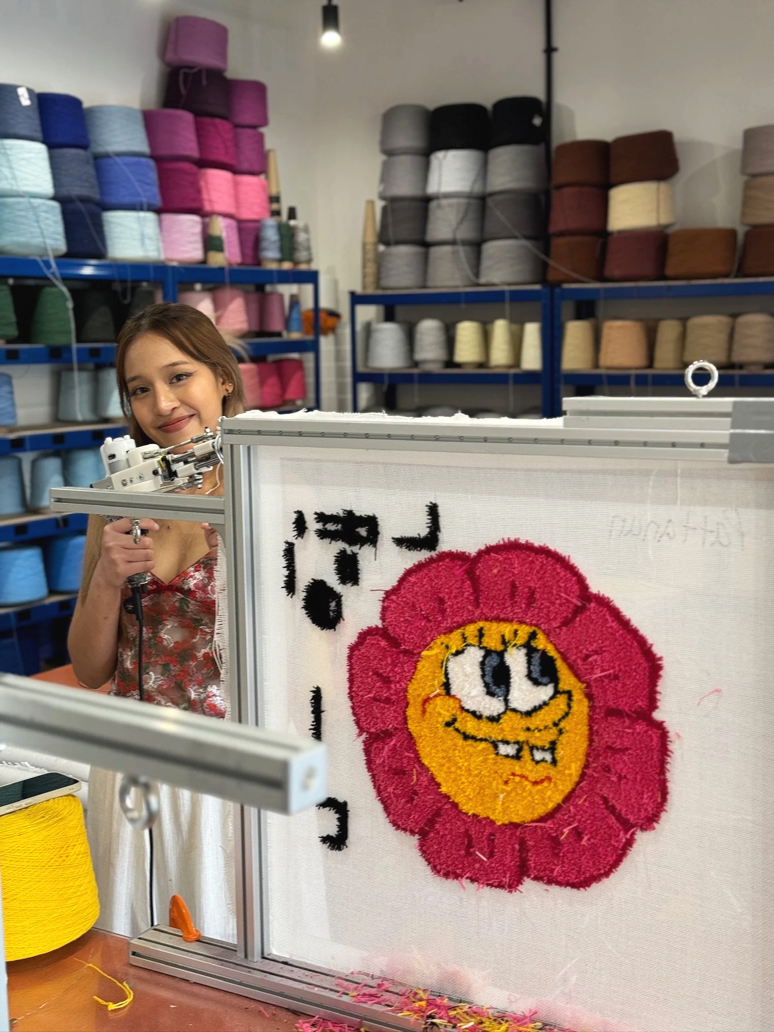 A smiling woman posing behind a floral embroidered fabric on a machine in a textile workshop with shelves of colorful yarn in the background.