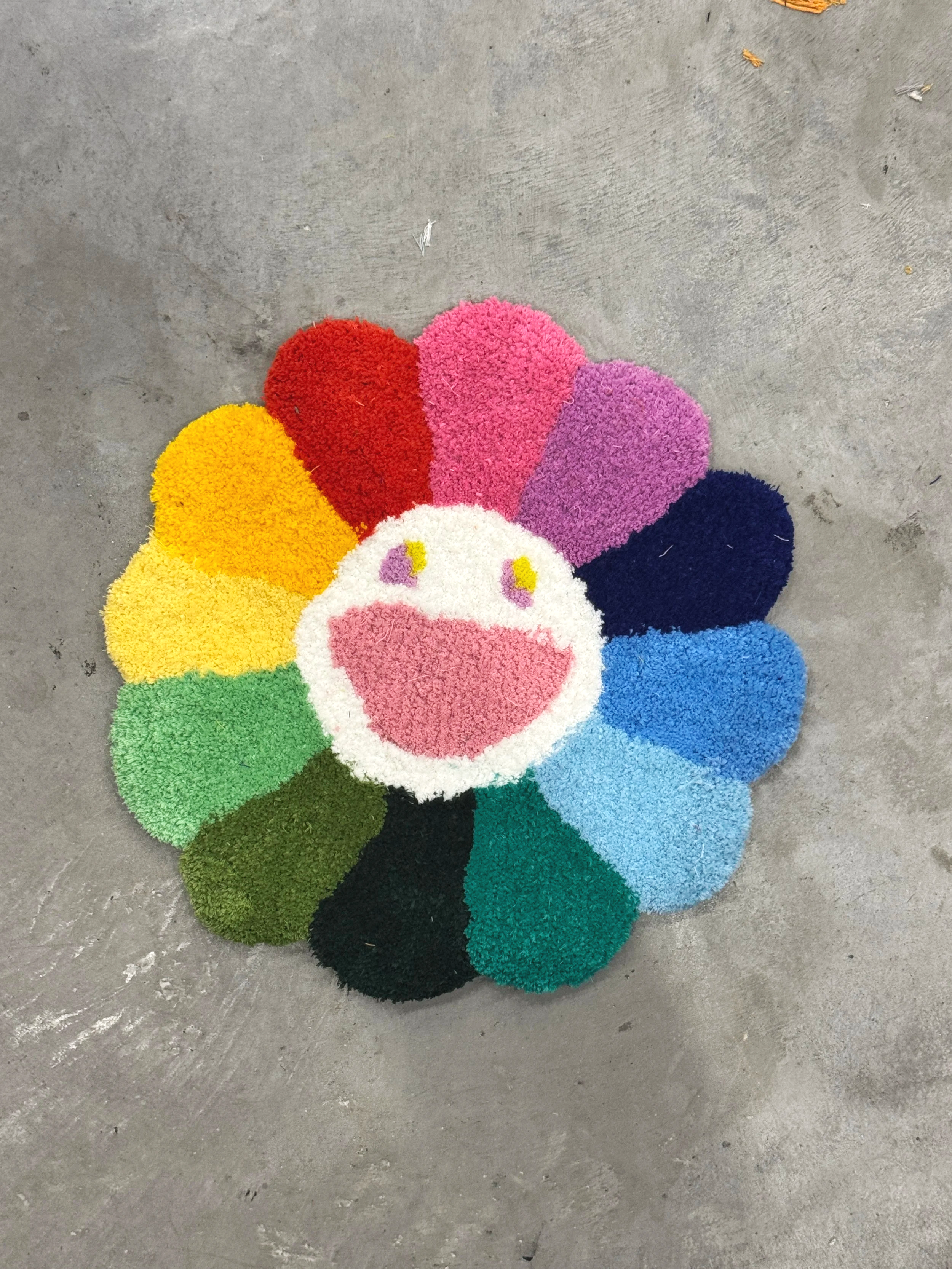 Colorful flower-shaped rug with a smiling face in the center, placed on a concrete floor.