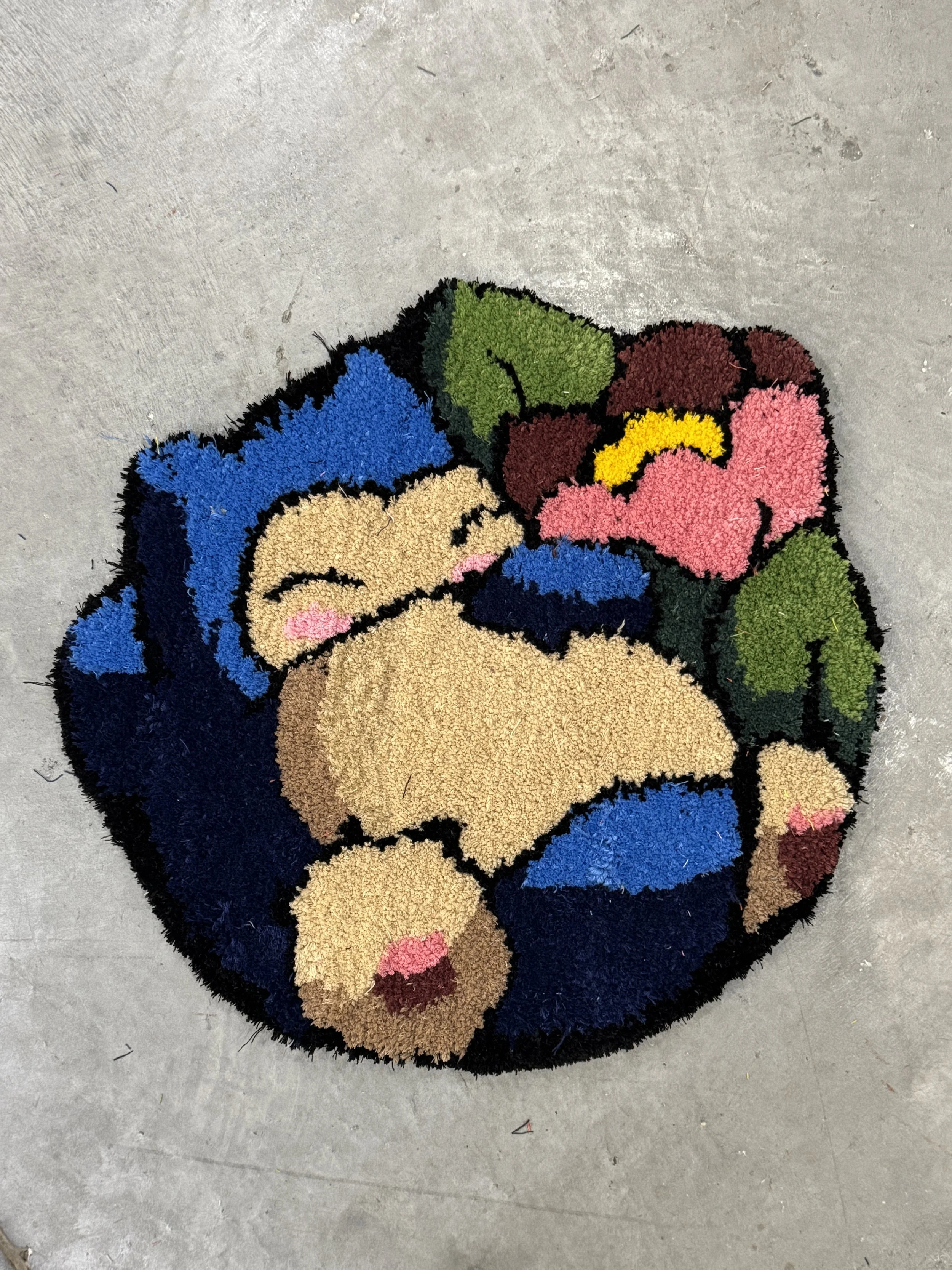Colorful doormat with a design of a cartoon character and flowers, featuring shades of blue, pink, green, yellow, beige, and brown on a concrete floor.