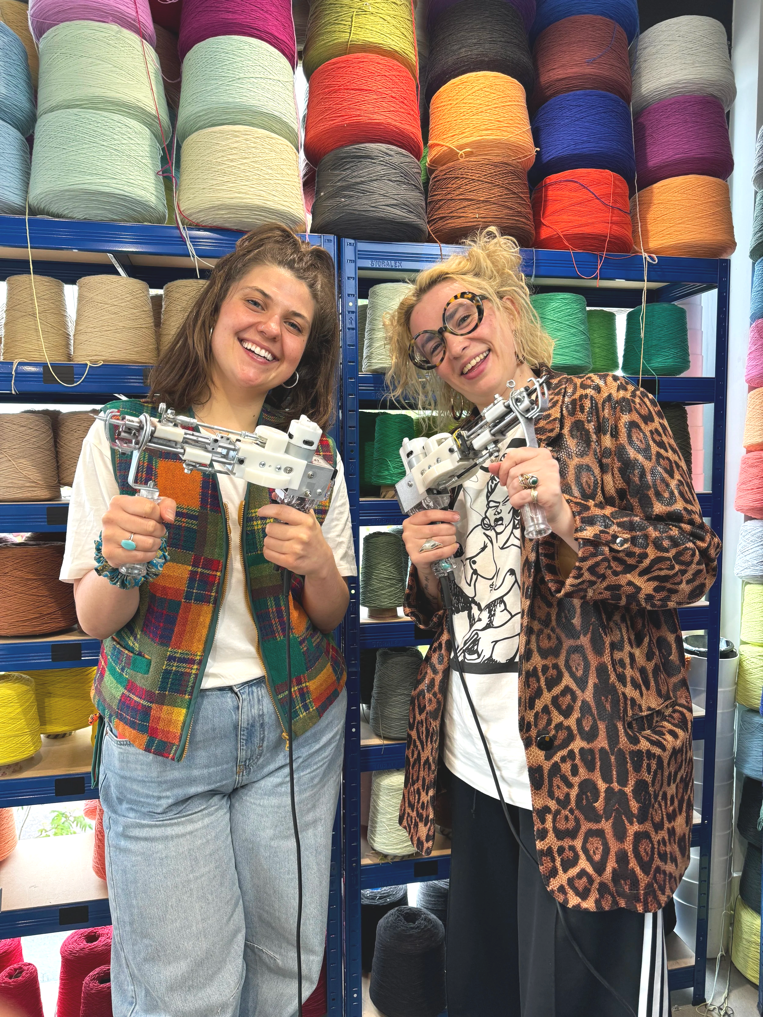 Two women smiling and holding sewing machines inside a yarn store with shelves of colorful yarn in the background.