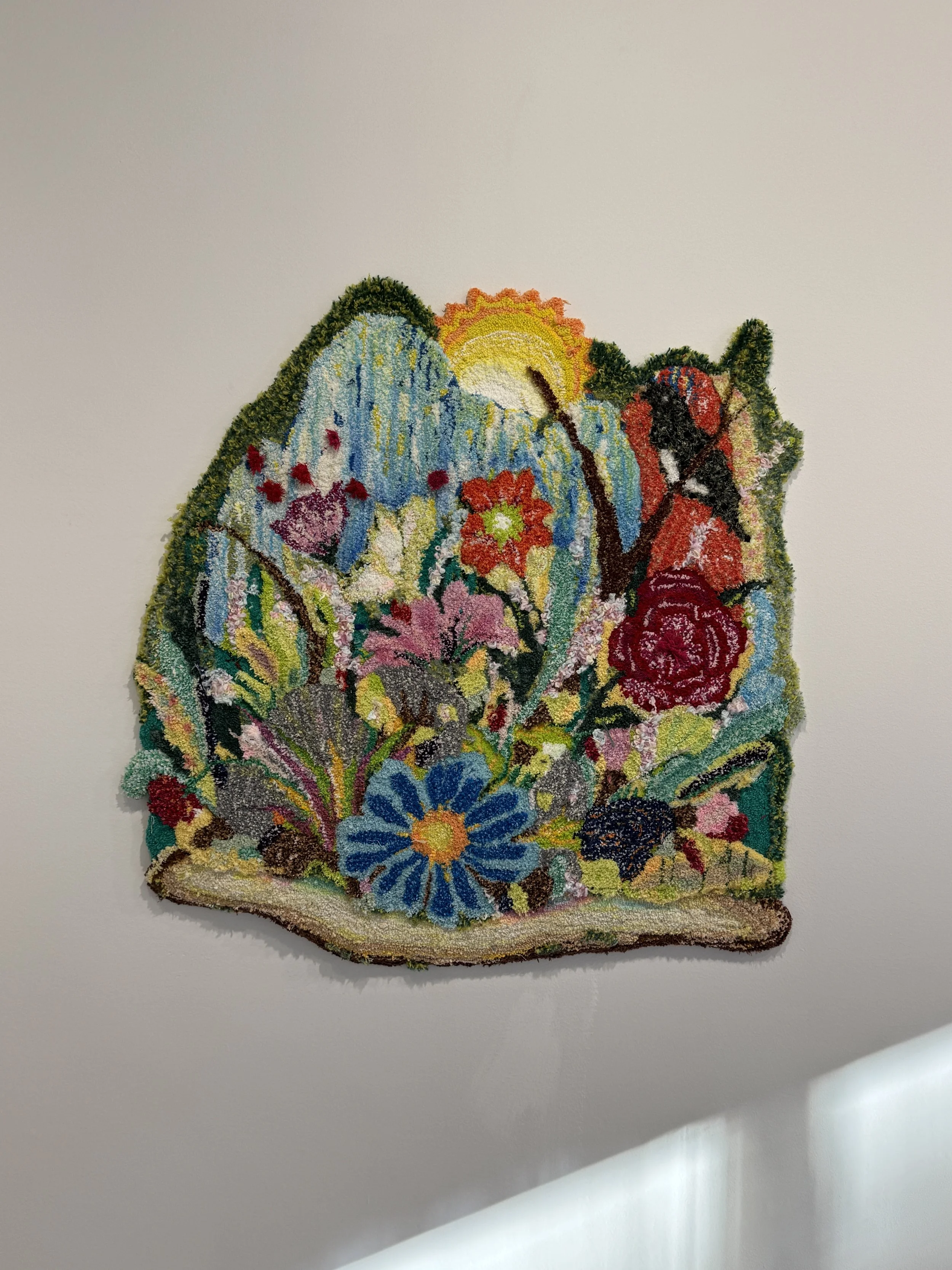 Colorful textile wall art featuring flowers, mountains, sun, and landscape elements on a plain wall.