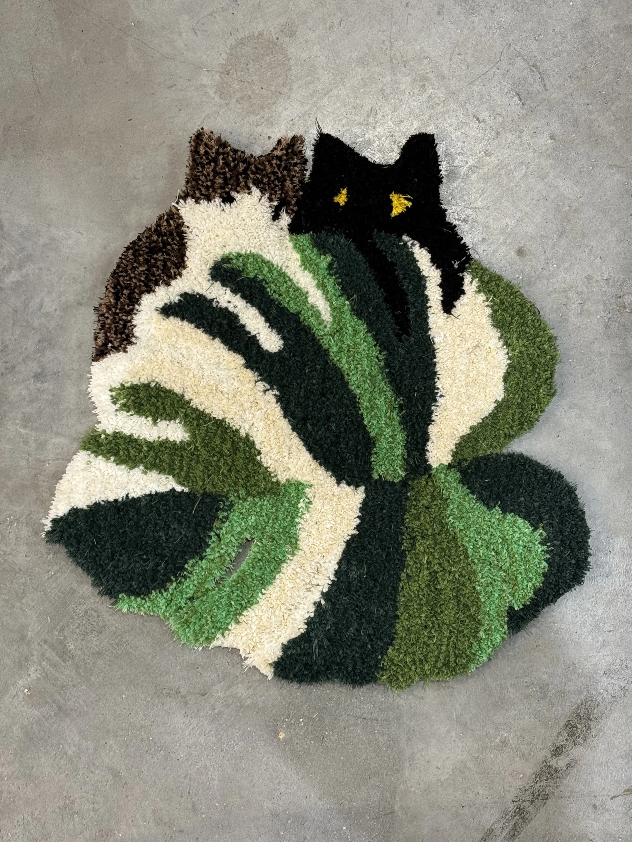 Colorful rug with a black cat face and green, white, black, and brown leaf pattern, placed on a concrete floor.