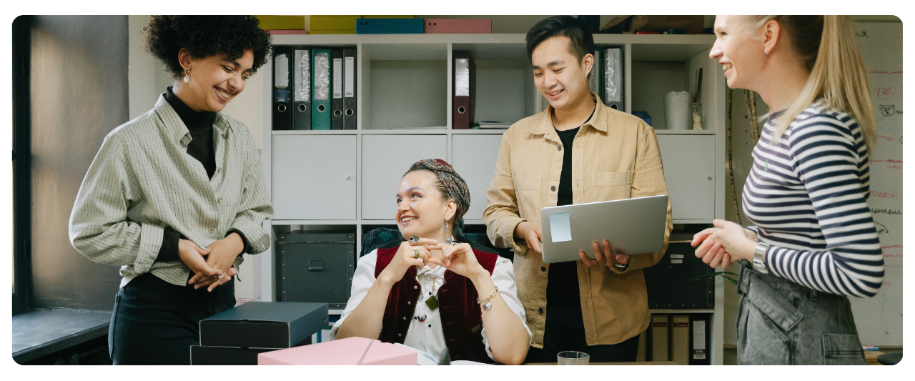 Four people in an office are smiling and talking, with shelves of binders and office supplies in the background.