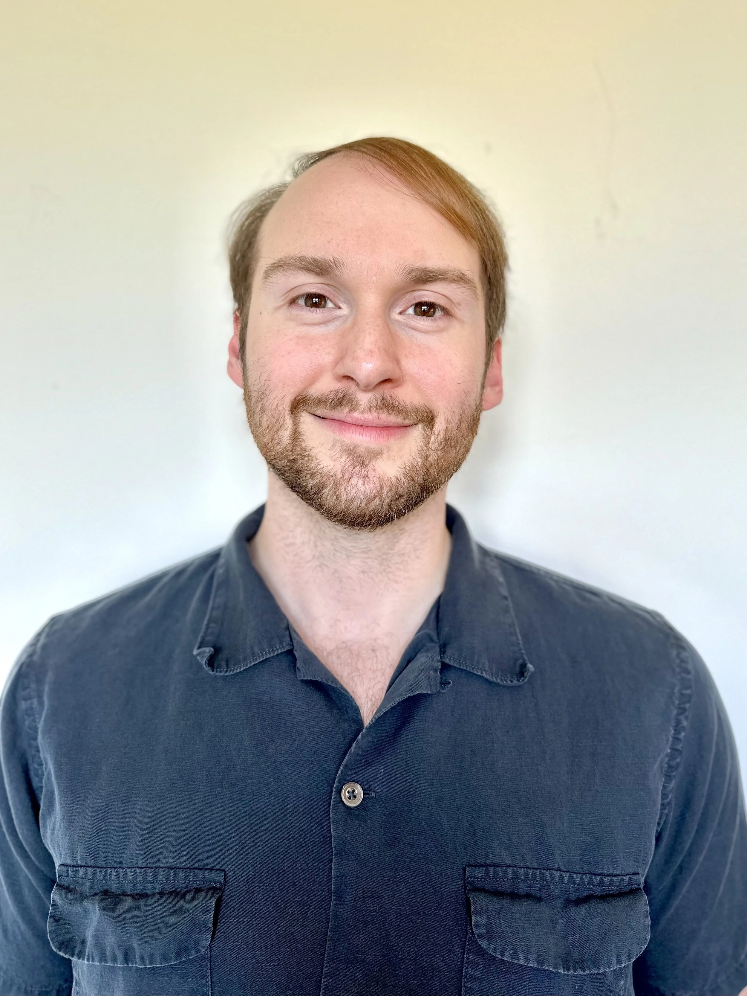 Portrait of a young man with short reddish-brown hair, light beard, wearing a dark button-up shirt, smiling against a plain light-colored background.