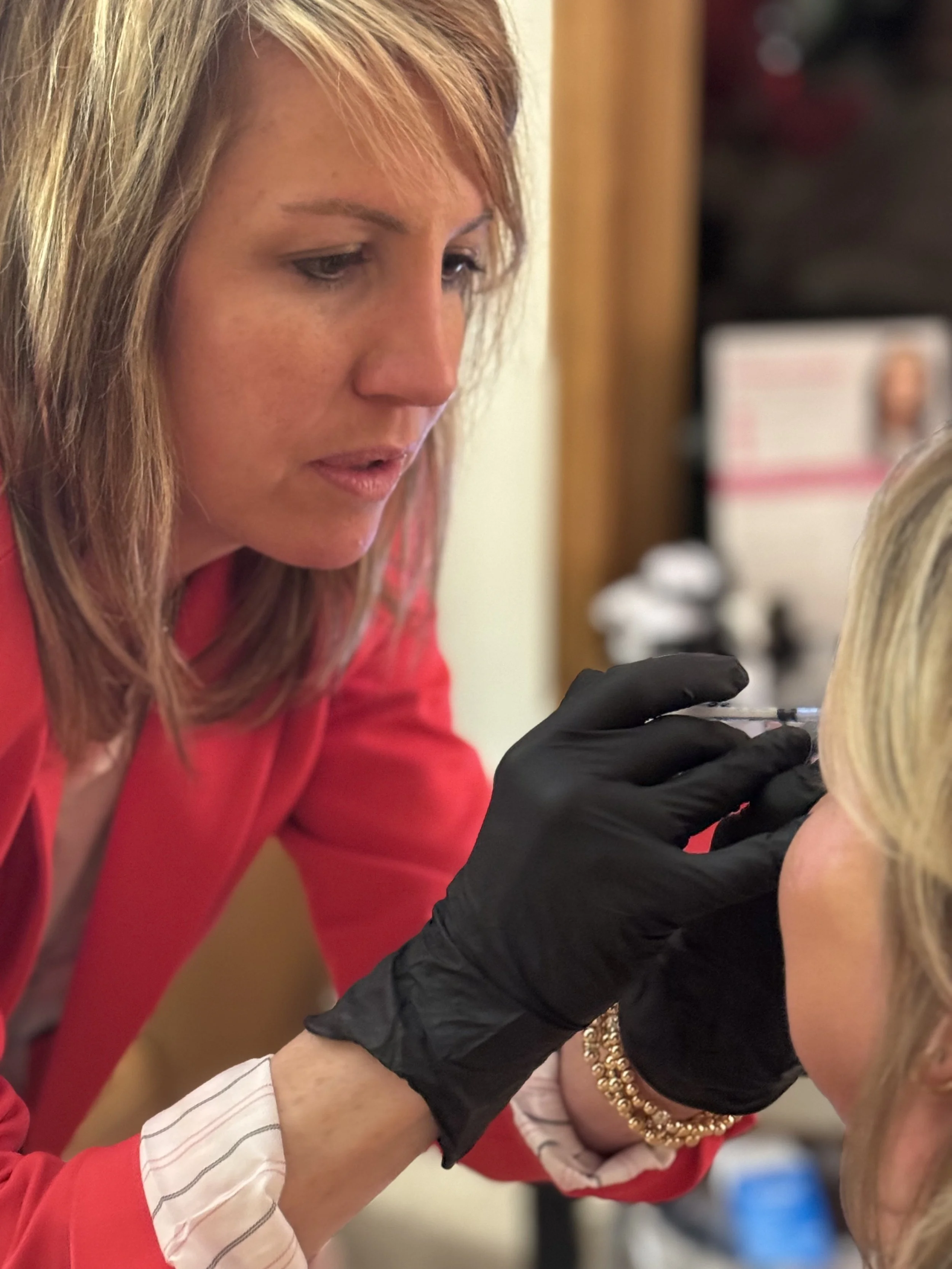 Woman in red coat performing cosmetic injection on another woman, wearing black gloves.