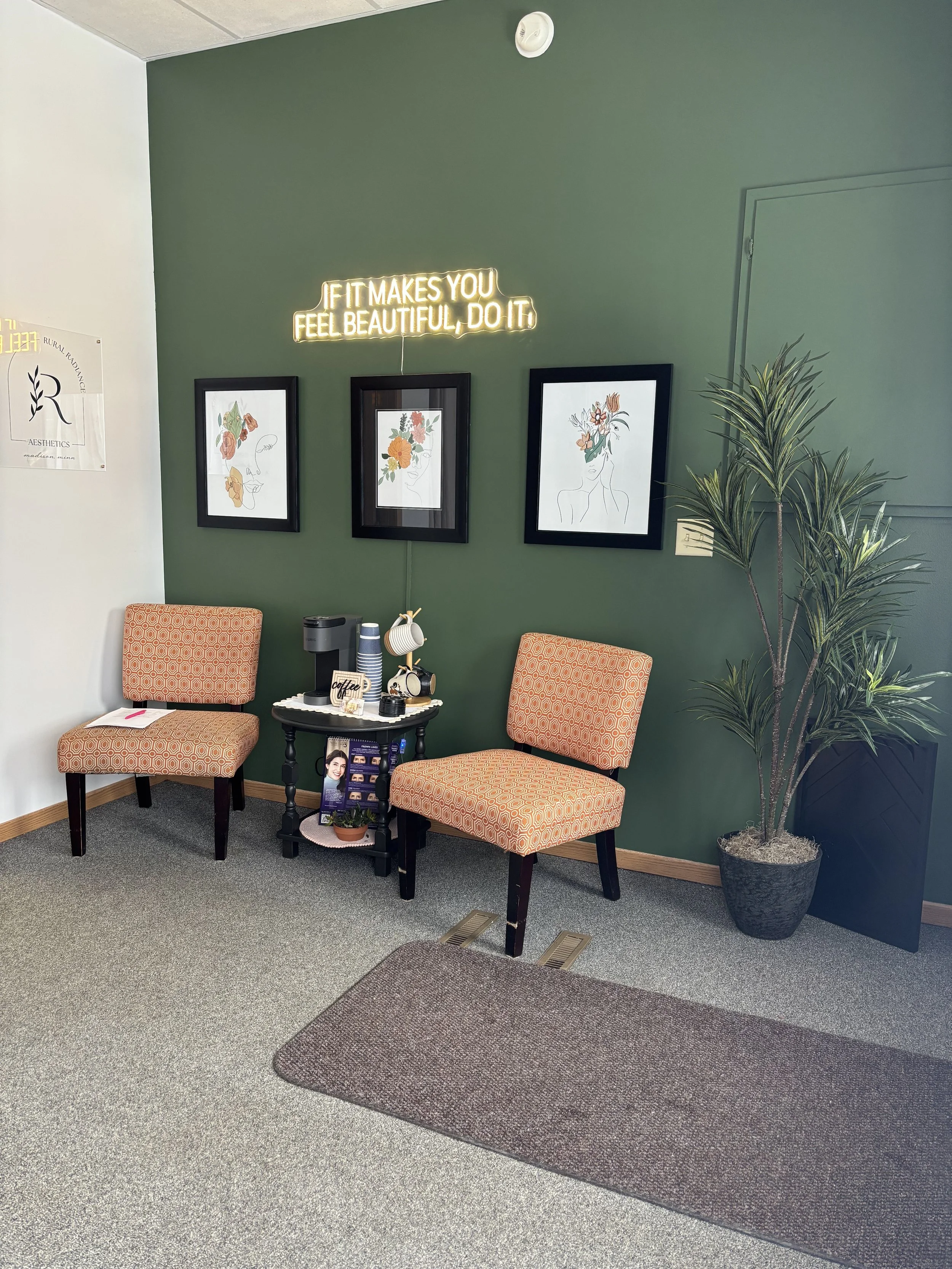 Stylish waiting area with two patterned chairs and a small table against a green wall, featuring three framed artworks. Neon sign above reads 'If it makes you feel beautiful, do it.' A plant is on the right, and there's a rug on the gray floor.