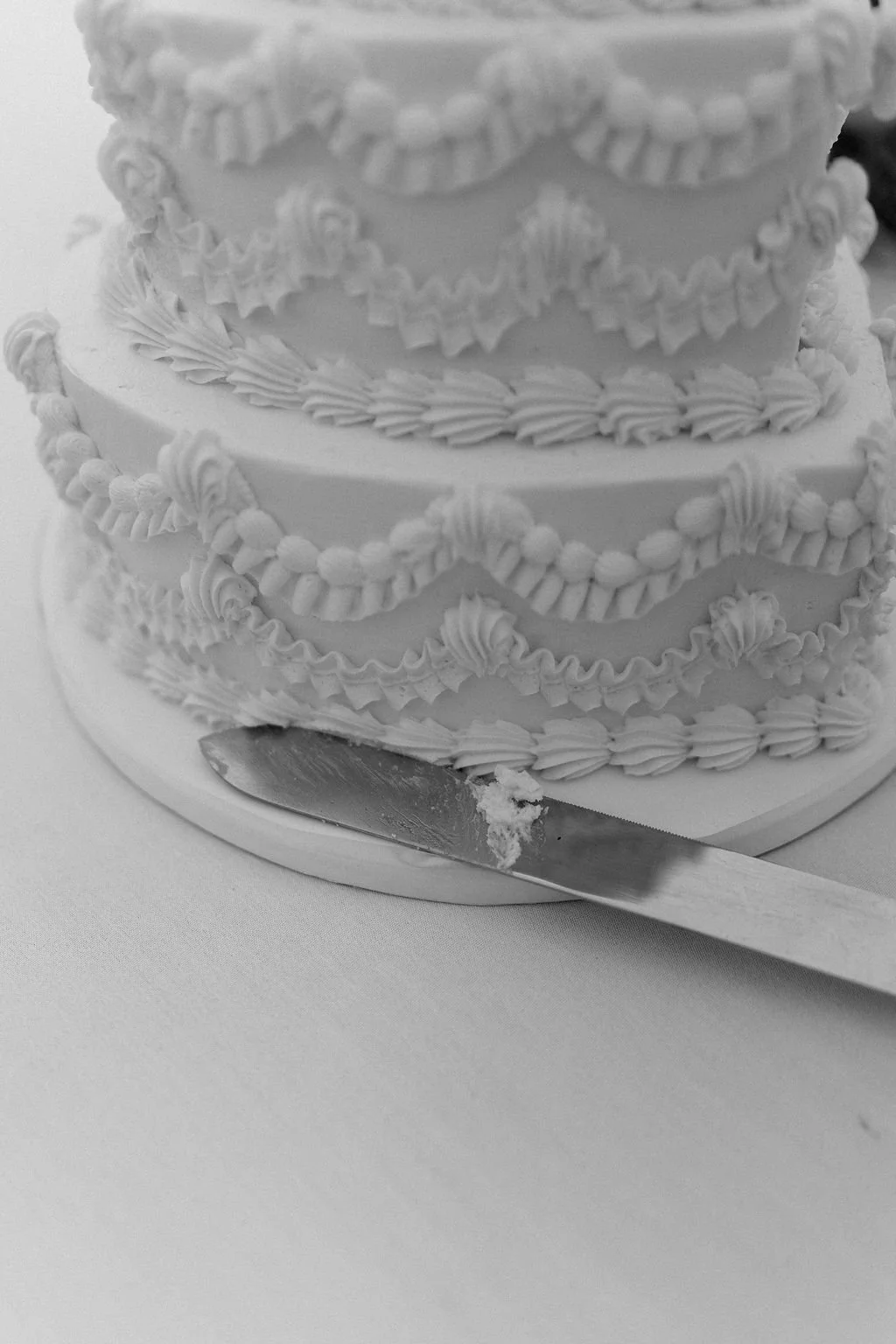 A decorative, multi-tiered wedding cake with intricate icing designs, placed on a white cake board. There is a serving knife with some cake remnants in the foreground.