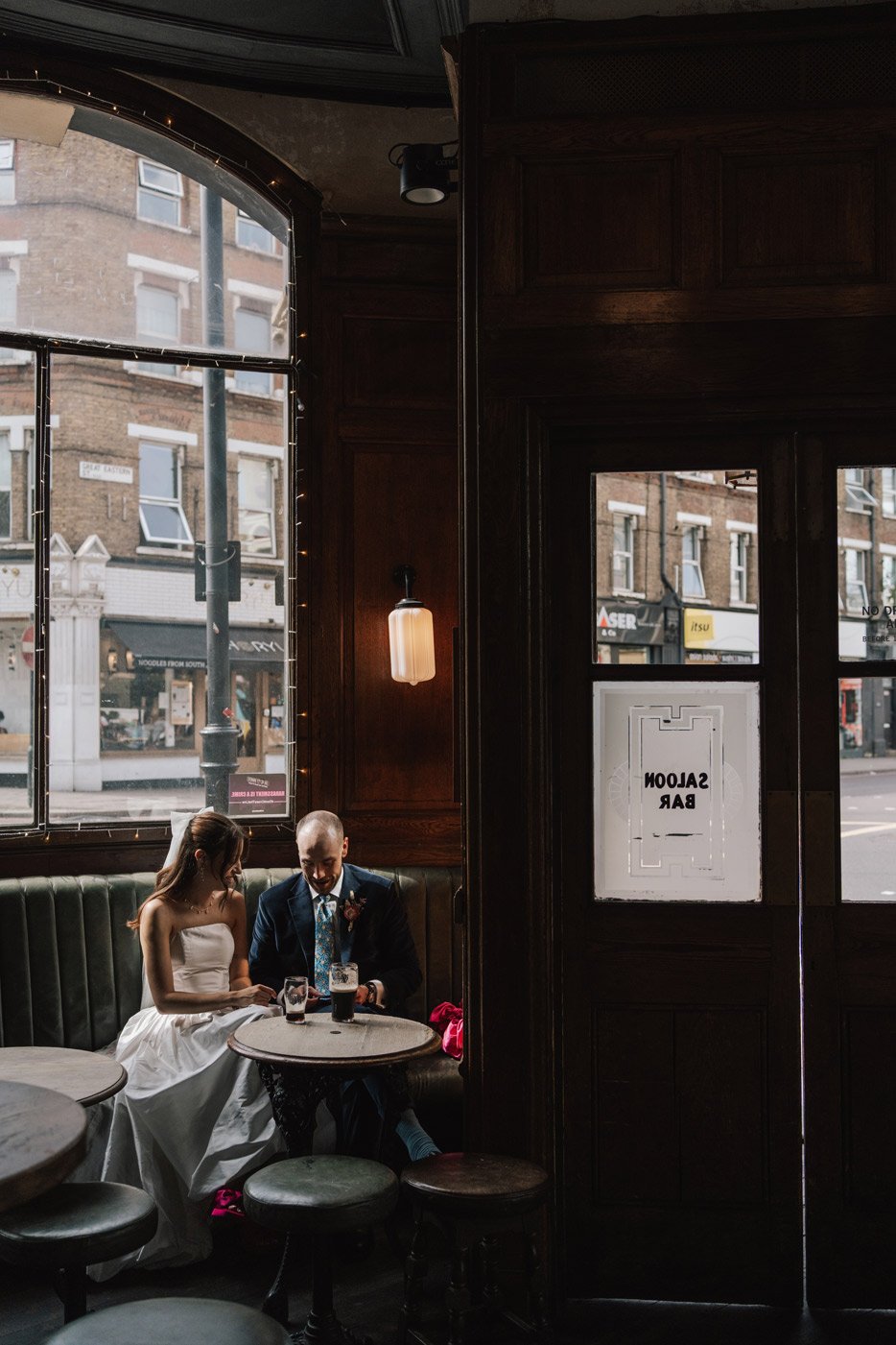 A bride and groom sit inside a dimly lit pub near a large window, sharing drinks and looking at a phone together, with a 'Saloon Bar' sign on the door