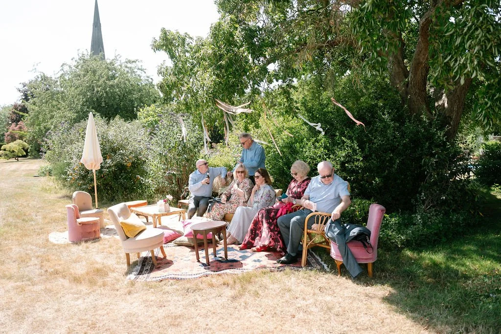 Group of people sitting on outdoor furniture in a garden, with trees and a church steeple visible in the background.
