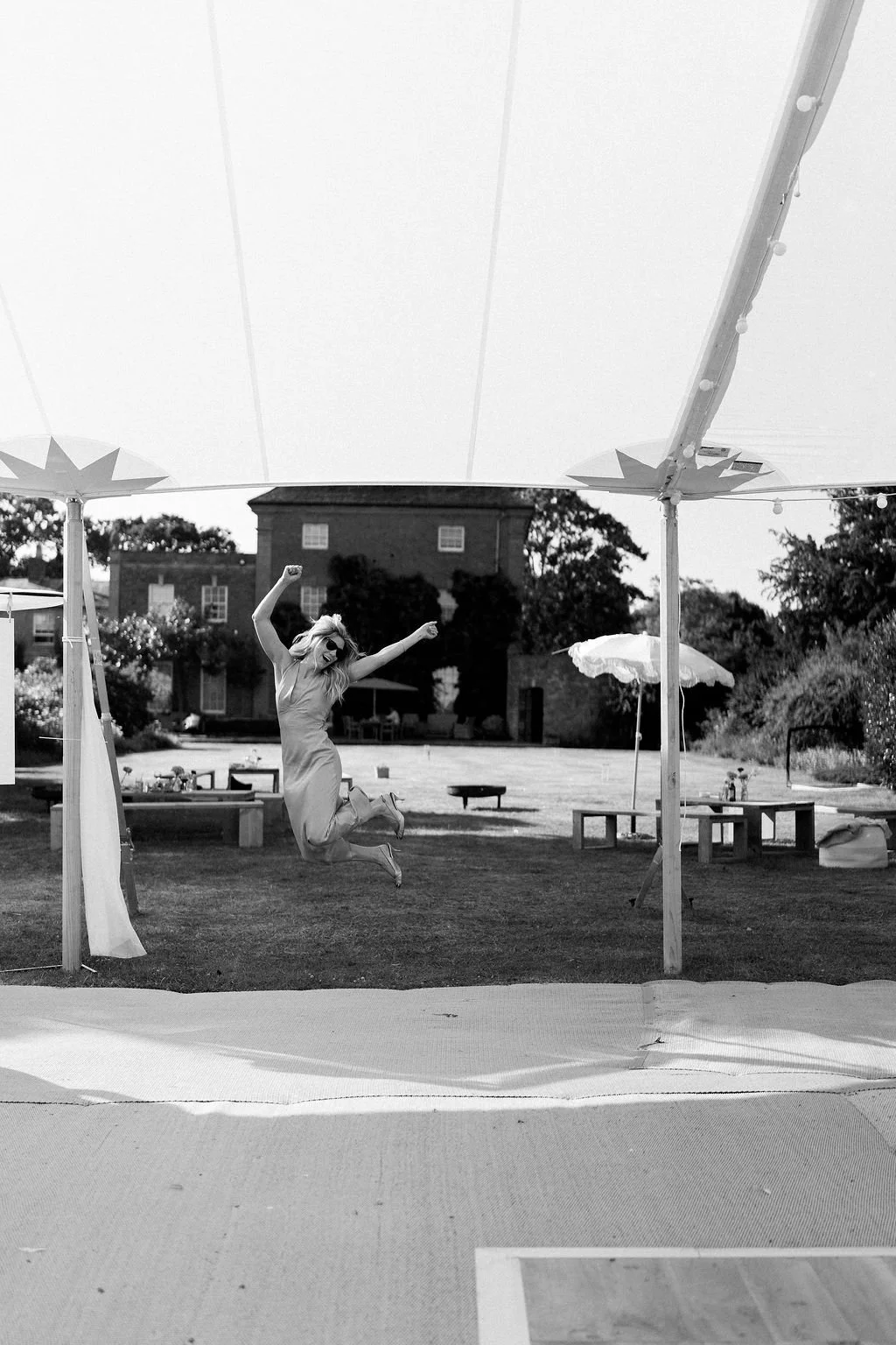 A woman jumping in the air under a large canopy at an outdoor event or party, with tables, chairs, and umbrellas in the background.