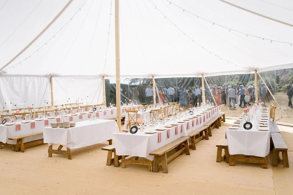 Long dining tables with white tablecloths, pink napkins, and floral centerpieces under a large white tent, with people outside mingling.